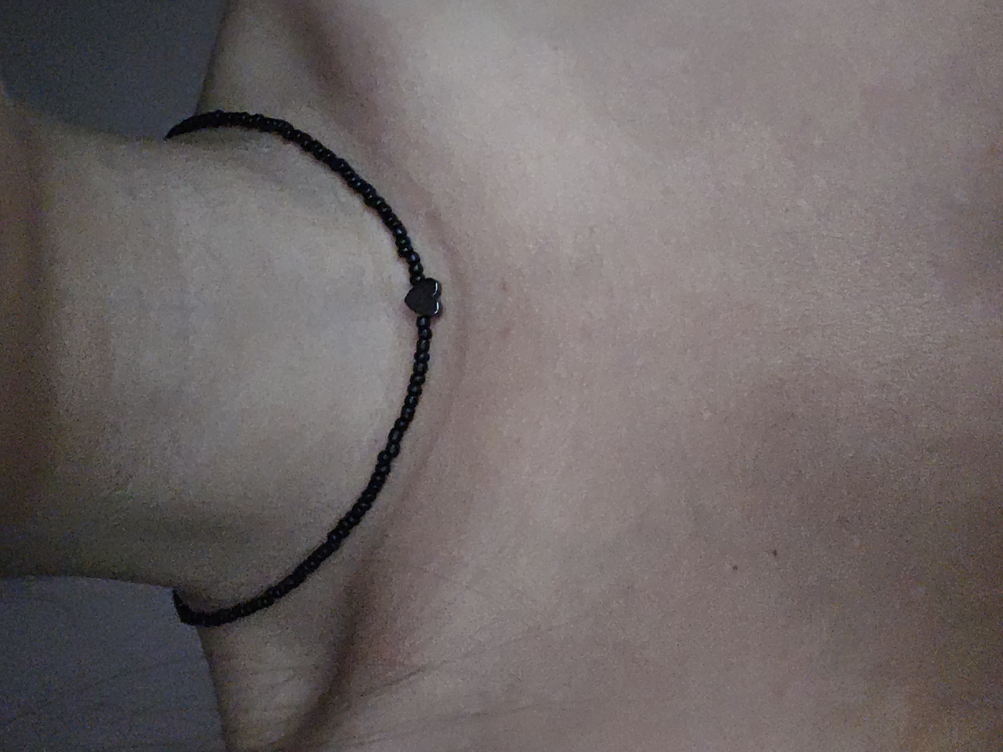 A close-up shot shows a person's neck and upper chest wearing a black beaded choker necklace with a small, dark heart pendant in the center. The background is a plain, textured wall in a muted gray color. The image does not provide any information about the city of Petropavlivska Borshchahivka, Ukraine.