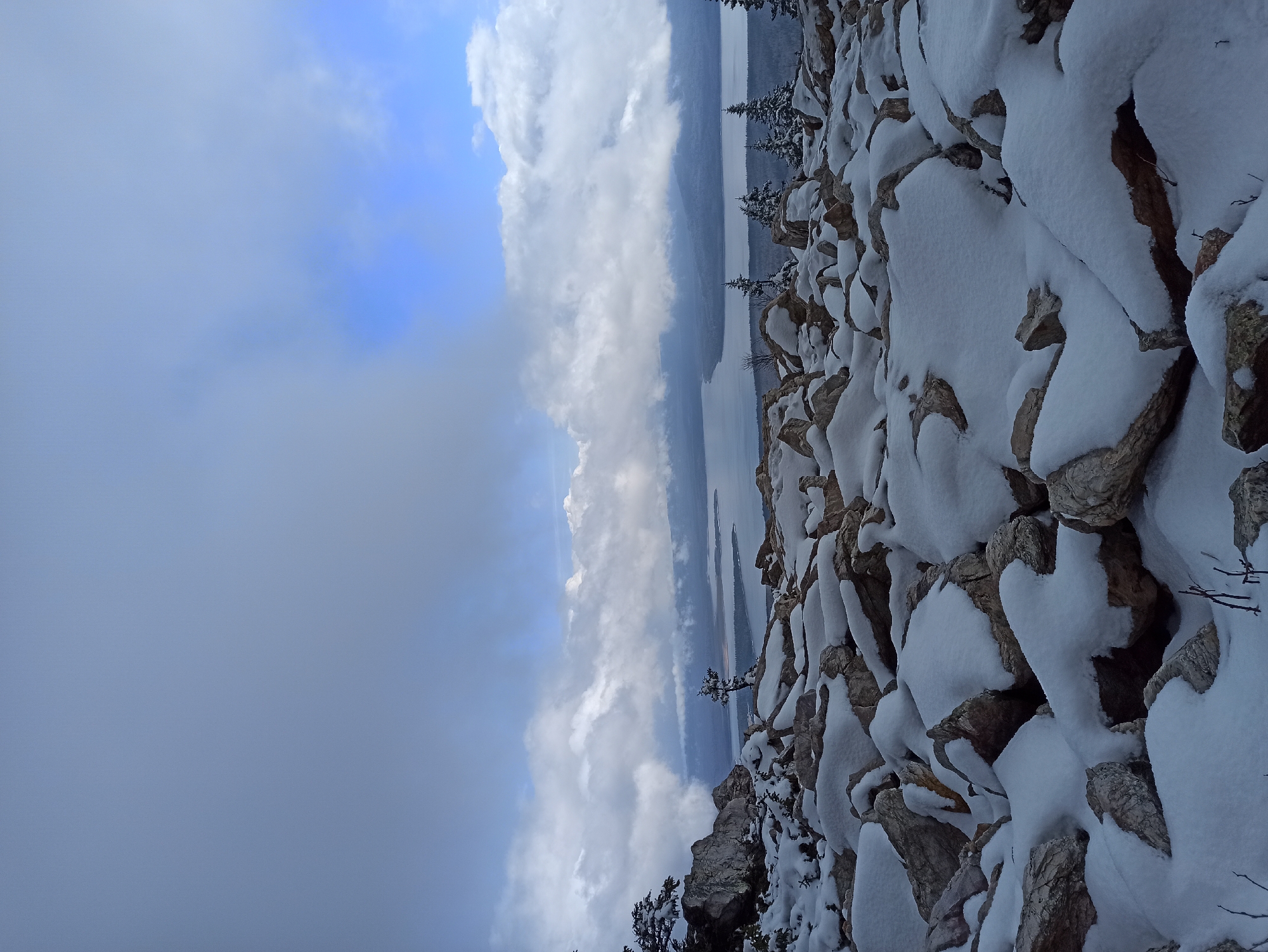 The image shows a rocky, snow-covered landscape. The scene includes rocks and ground covered in snow. A body of water, possibly a lake or river, stretches across the middle ground, partially obscured by a layer of fog. The sky is covered by clouds. The image could be in Ziuratkul', Russia.