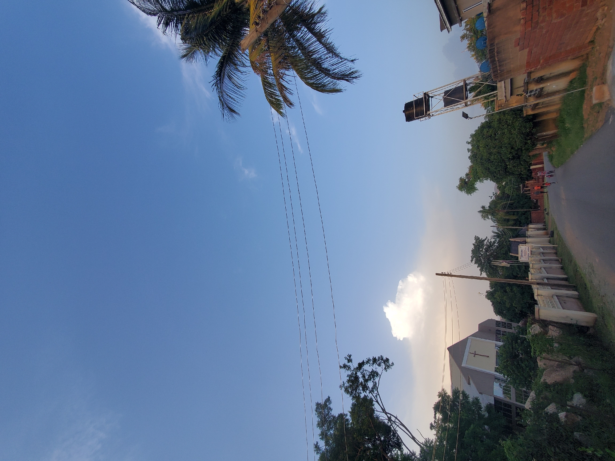 The image depicts a street scene in Jos, Nigeria, under a clear blue sky with scattered clouds. On the left side of the street, a modern church building with a cross visible on its facade is surrounded by lush green trees. A concrete fence with decorative elements runs along the front of the church property. The road is paved and appears to be a residential or community area.
To the right of the road, a tall water tank structure is prominently featured, leaning slightly. Adjacent to the water tank is a brick wall. Further down the road, several people, including children, are walking. The environment is characterized by vegetation, including trees and grass, alongside man-made structures. Overhead, power lines are visible, strung across the frame. The overall impression is of a daytime scene in a developing urban or suburban area.
