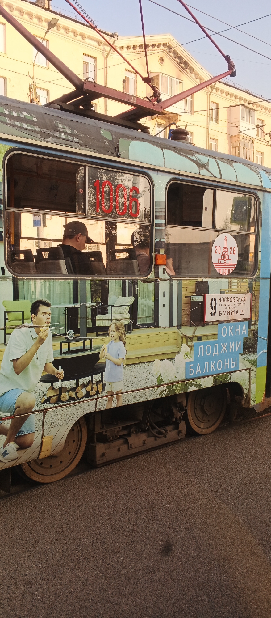 This is an exterior, eye-level, medium shot of a tram decorated with a mural on a street in Izhevsk, Russia. The tram has a teal and blue painted exterior with images of people blowing bubbles, wooden furniture, and text. The text on the tram reads "1006" in red numerals on the window, "20 26" with a graphic of a building on a circular emblem, and three blue blocks with white text that say "ОКНА", "ЛОДЖИИ", and "БАЛКОНЫ". A white sign with a black border has a black number "9" on it and smaller text that reads "МОСКОВСКАЯ И.К. МАРКСА - К. ЛИБКНЕХТА И. СЕРГЕЯ БУММАШ". Inside the tram, two passengers are visible through the windows. The tram is on a paved street, and a building with yellow facade and ornate decorations is visible in the background. The tram's pantograph is connected to overhead wires. The tram's wheels and undercarriage are visible at the bottom of the image.