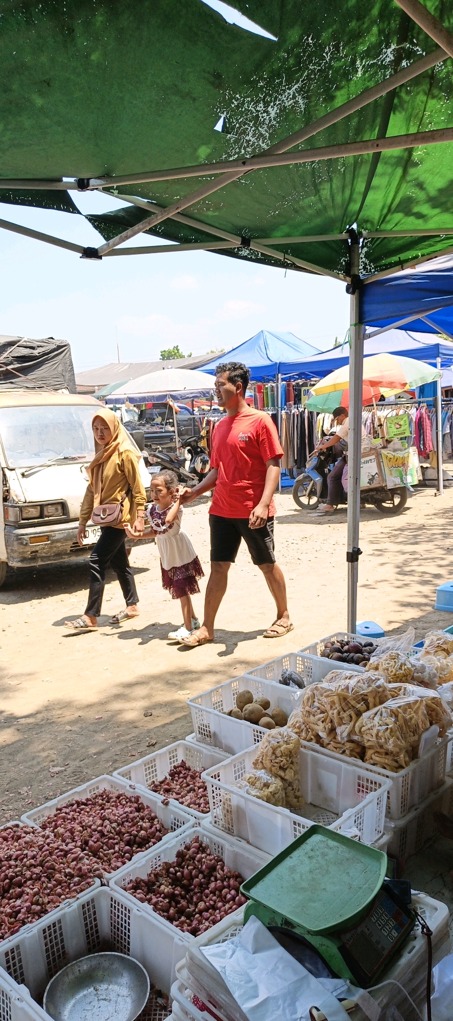 In Jatinom, Indonesia, a family of three, a father, mother, and daughter, are walking through an outdoor market. The mother, wearing a tan hijab and a patterned dress, walks to the left, while the father, in a red t-shirt and black shorts, holds his daughter's hand on the right. The daughter, in a white dress with purple trim, walks between them. In the foreground, baskets of red onions and other goods are displayed for sale. A scale with a green tray sits on top of a stack of baskets. In the background, market stalls with colorful awnings are set up, with a van and motorcycles visible. The scene appears to be bustling with activity under the bright, sunny sky.