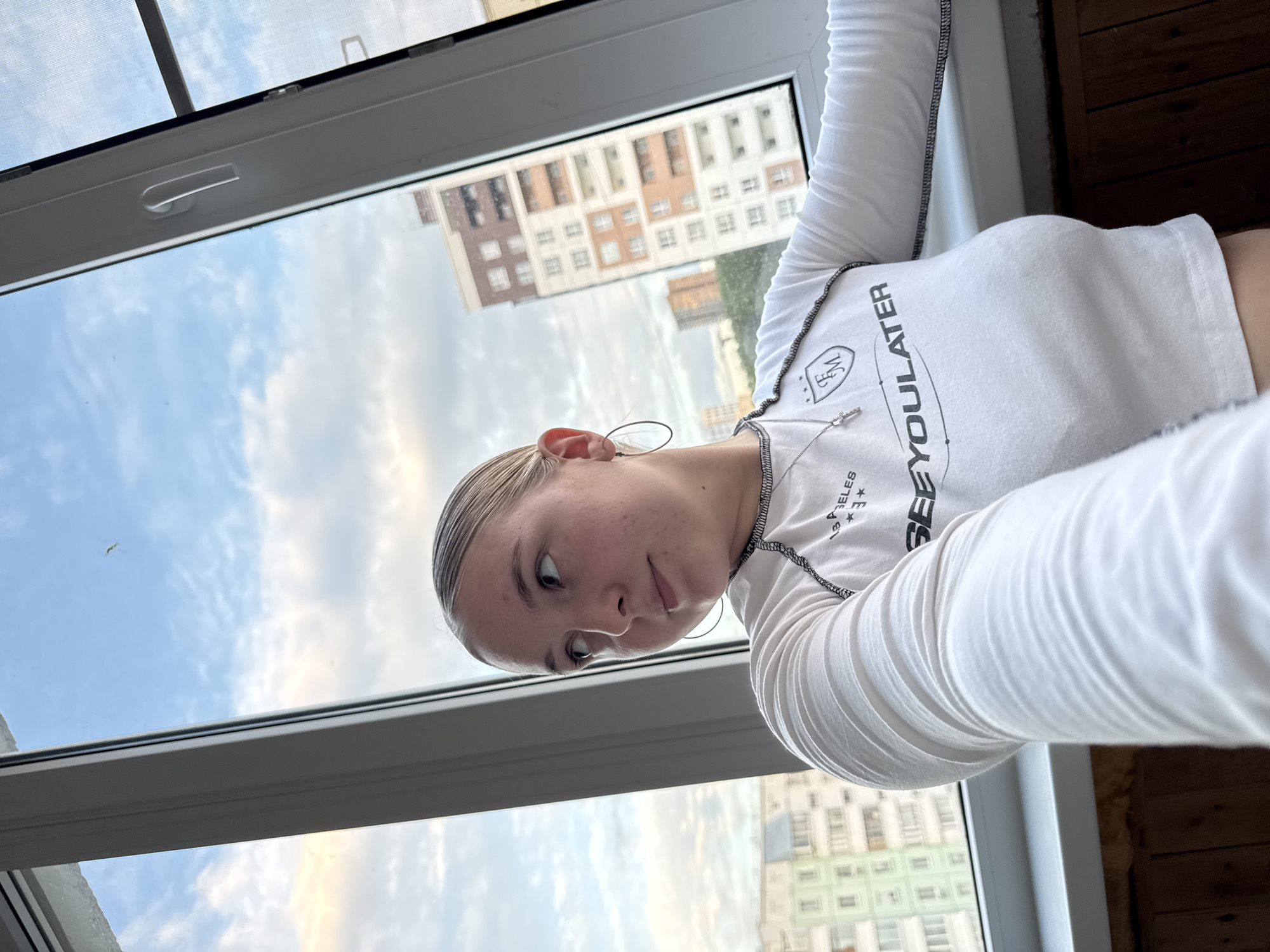 A young woman with blonde hair pulled back in a bun is standing near a window, looking out at an urban landscape. She is wearing a white long-sleeved cropped shirt with black trim and text that reads "SEE YOO LATER". She also has a silver necklace with a cross pendant. The background shows apartment buildings and a cloudy sky. The window has a white frame and a handle. The lower part of the image shows a wooden surface. This scene is located in the city of Perm, Russia.