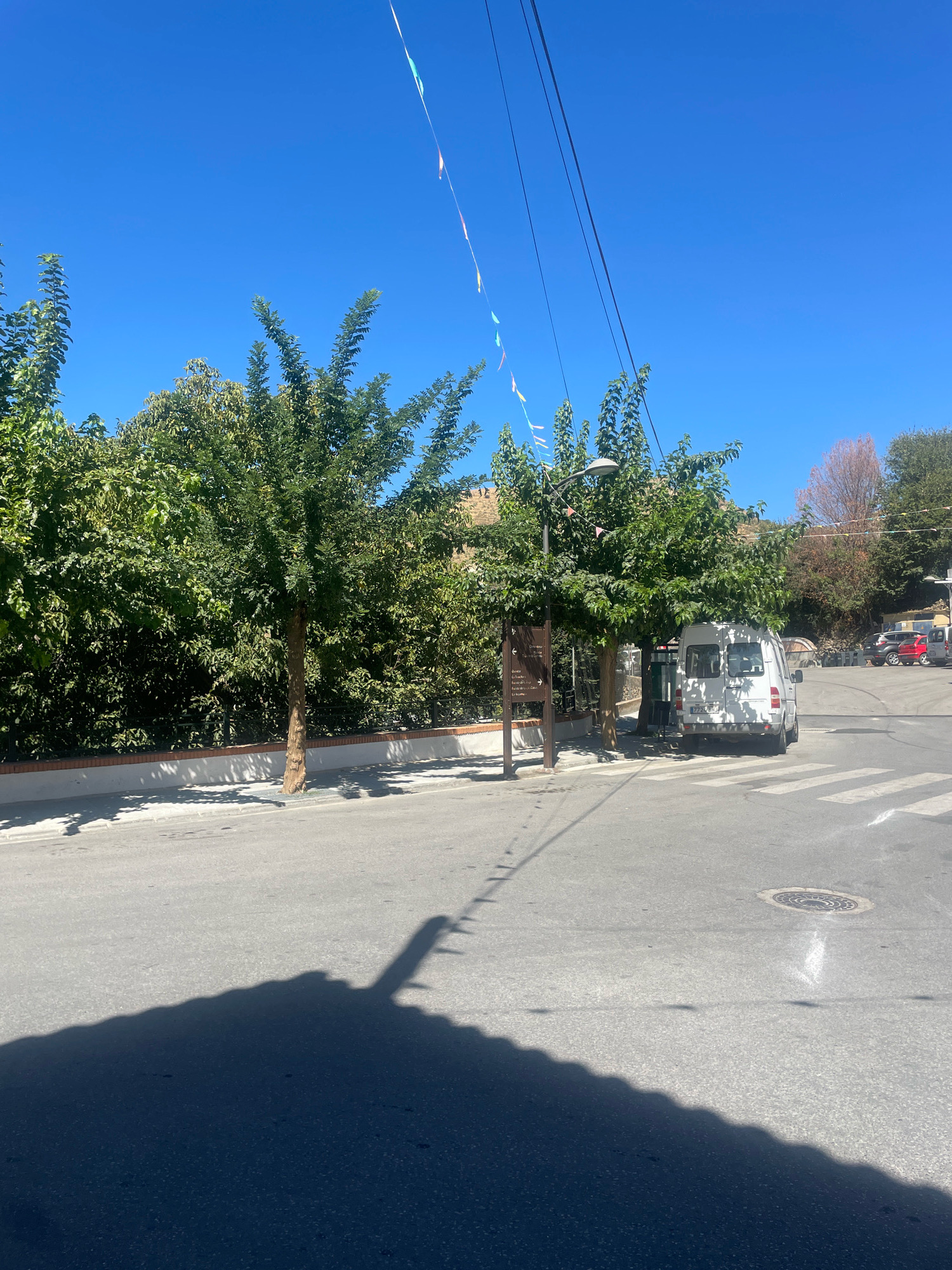 The image depicts a street scene in Güéjar Sierra, Spain, under a clear blue sky. In the foreground, a dark shadow of an unseen object, possibly a building or a large structure with a serrated edge, covers a significant portion of the asphalt road. The road itself is gray and appears to be in good condition, with a pedestrian crossing marked by white stripes. A manhole cover is visible on the right side of the road.
Further into the scene, a white van with a license plate reading "2772 BRJ" is parked on the right side of the street, partially obscuring a pedestrian crossing. To the left of the van, two trees with green foliage stand on the sidewalk. The left tree is taller and more slender, with a visible brown trunk. The right tree is wider and more spread out. A street lamp is mounted on a pole situated between the two trees. Next to the left tree, a dark brown wooden signpost with text is visible.
Behind the trees and the sidewalk, a brick wall topped with a low white border runs along the property line. Beyond the wall, dense green foliage from trees and bushes creates a thick, natural barrier. In the background, several buildings with reddish-brown roofs are nestled on a hillside, interspersed with more trees. Some of these trees show autumnal colors, with a prominent tree exhibiting reddish-brown foliage. Decorative triangular flags, in various colors including white, green, and orange, are strung across the scene at the top, suggesting a festive occasion. Power lines run diagonally across the upper portion of the image. The overall impression is a sunny day in a town with a mix of natural greenery and urban infrastructure.