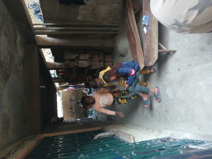 The scene depicts a narrow, covered concrete passageway in the city of Mgbidi, Nigeria. The passage features a concrete ceiling with an exposed light bulb fixture and a concrete floor. The left wall is partially obscured by a green metal security gate. The right wall is solid concrete, with vertical elements that could be rebar or conduits, and stacks of light-colored, possibly wooden or concrete, forms are visible further back.

Four individuals are present in the scene, plus a fifth person whose back and shoulder are visible in the lower right foreground, presumably taking the photograph.

In the mid-ground, a young female, possibly a teenager, with braided hair, sits on a rustic wooden bench on the right side of the passage. She wears a dark blue long-sleeved top with red and white stripes, dark green pants, and grey sandals, and is focused on a white smartphone held in her hands. Directly in front of her is another wooden bench, on which a small blue item is placed.

Standing near the seated female are two younger children. To her right, a barefoot child in a yellow top and brown pants stands, observing the seated female. To the left and slightly behind, another child in an orange top and camouflage-patterned pants is captured in mid-stride, wearing dark sandals and looking towards the left.

In the distant background, at the end of the passageway, another individual is seated or crouched on the ground, with their features indistinct. Various items, including a blue container and draped fabrics, are visible in this area.