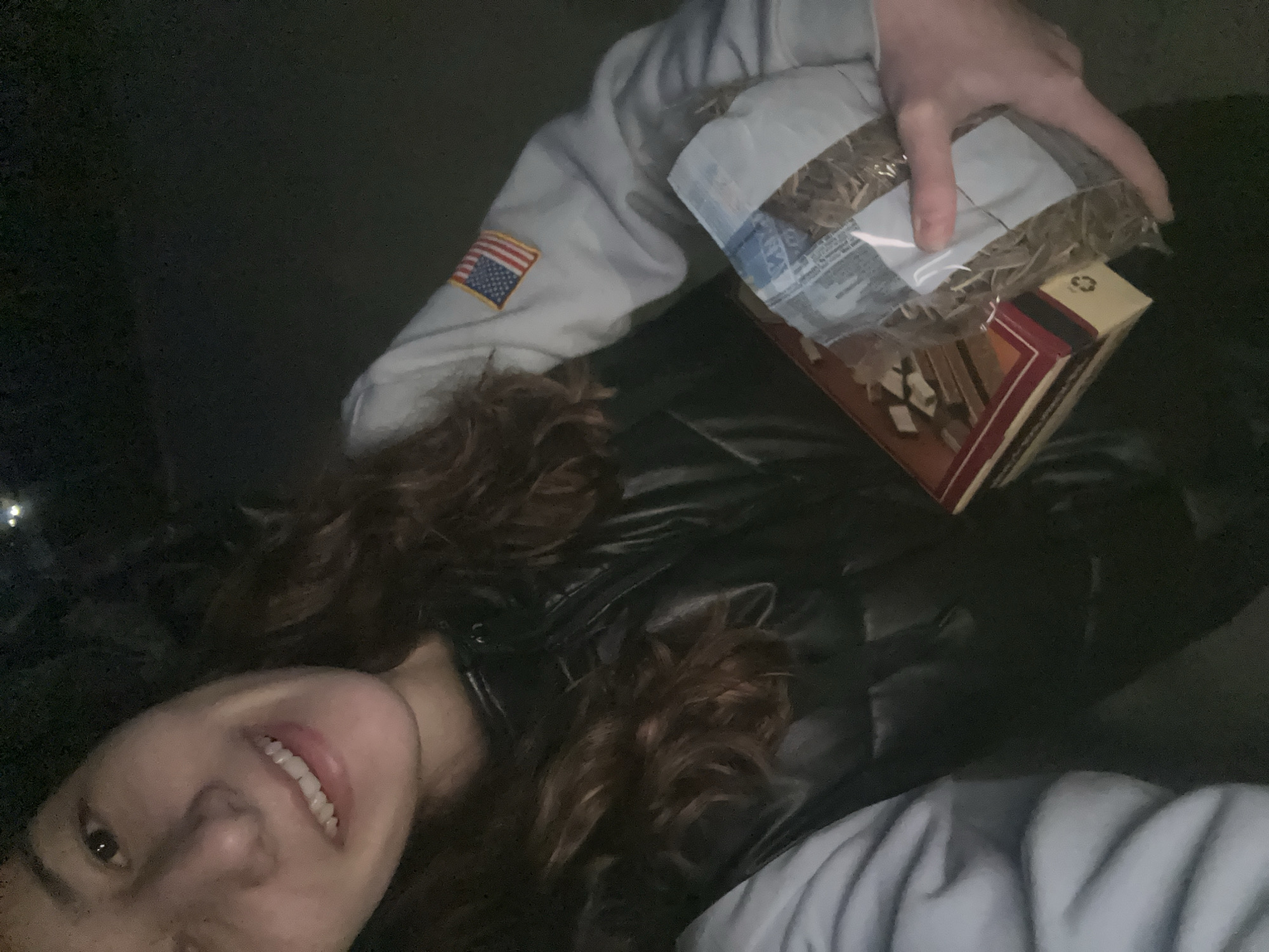 A young woman with long, curly brown hair and a fair complexion is holding a bag of hay and a rectangular box of wood kindling. She is wearing a dark, possibly leather, jacket and a light gray sweatshirt with an American flag patch on the sleeve. The background is dark, suggesting it is nighttime. The location is unknown and cannot be determined.