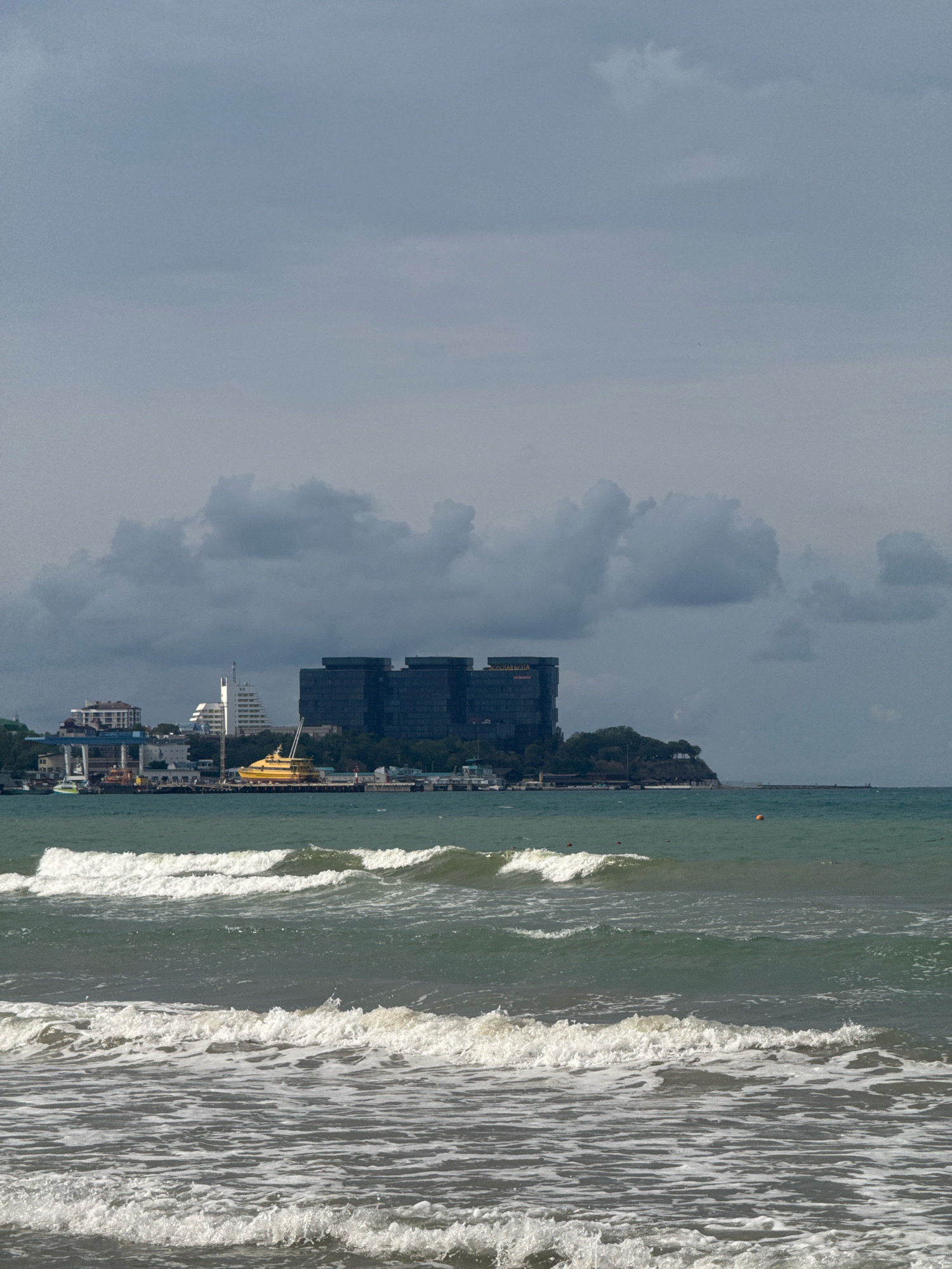 A horizontal, eye-level shot depicts a view of the Black Sea shoreline in Anapa, Russia, under a cloudy sky. In the foreground, three rows of gentle waves crest and break, creating white foam that dissipates into the darker green water. The mid-ground showcases a developed coastline with buildings and infrastructure. Prominently, a large, dark, modern building complex with many windows dominates the center-right of the horizon. To its left, a bright yellow vessel, resembling a catamaran or ferry, is docked. A white, slender structure, possibly a lighthouse or a modern art installation, stands near other buildings in the mid-left. A breakwater or pier extends into the sea on the right, with a single orange buoy visible in the water. The background is filled with a thick, grey, cloudy sky. The scene is devoid of any visible people.