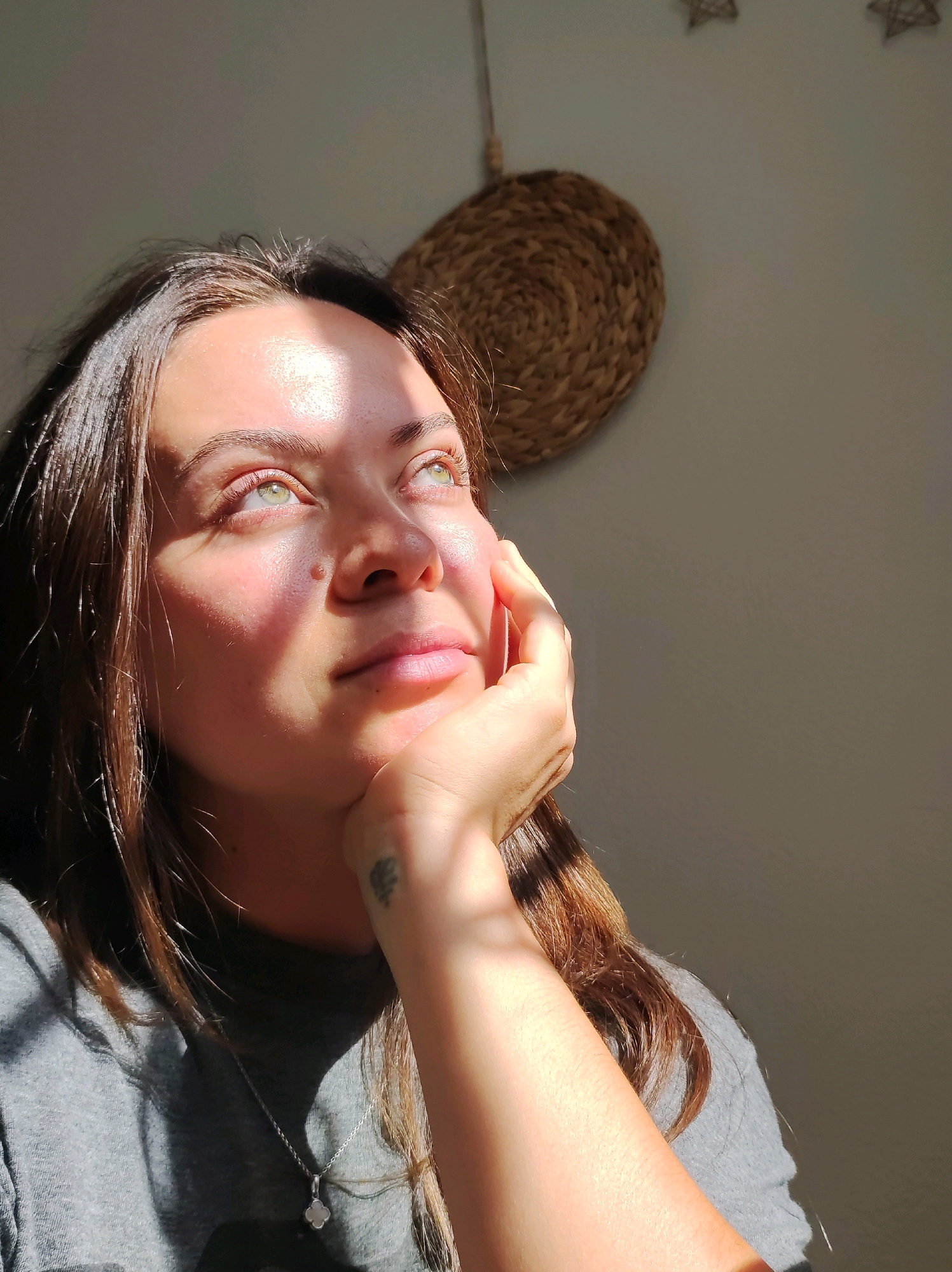 The image shows a close-up of a woman with brown hair and green eyes, looking upwards and to the right. She is resting her chin on her hand, and there is a subtle tattoo on her wrist. Sunlight is illuminating her face and casting shadows. She is wearing a grey t-shirt and a silver pendant. In the background, a woven decorative item is hanging on a wall. The overall lighting suggests it is daytime, with natural light coming from a window. There is no information in the image to indicate the location is the city of Chaikovsky, Russia.
