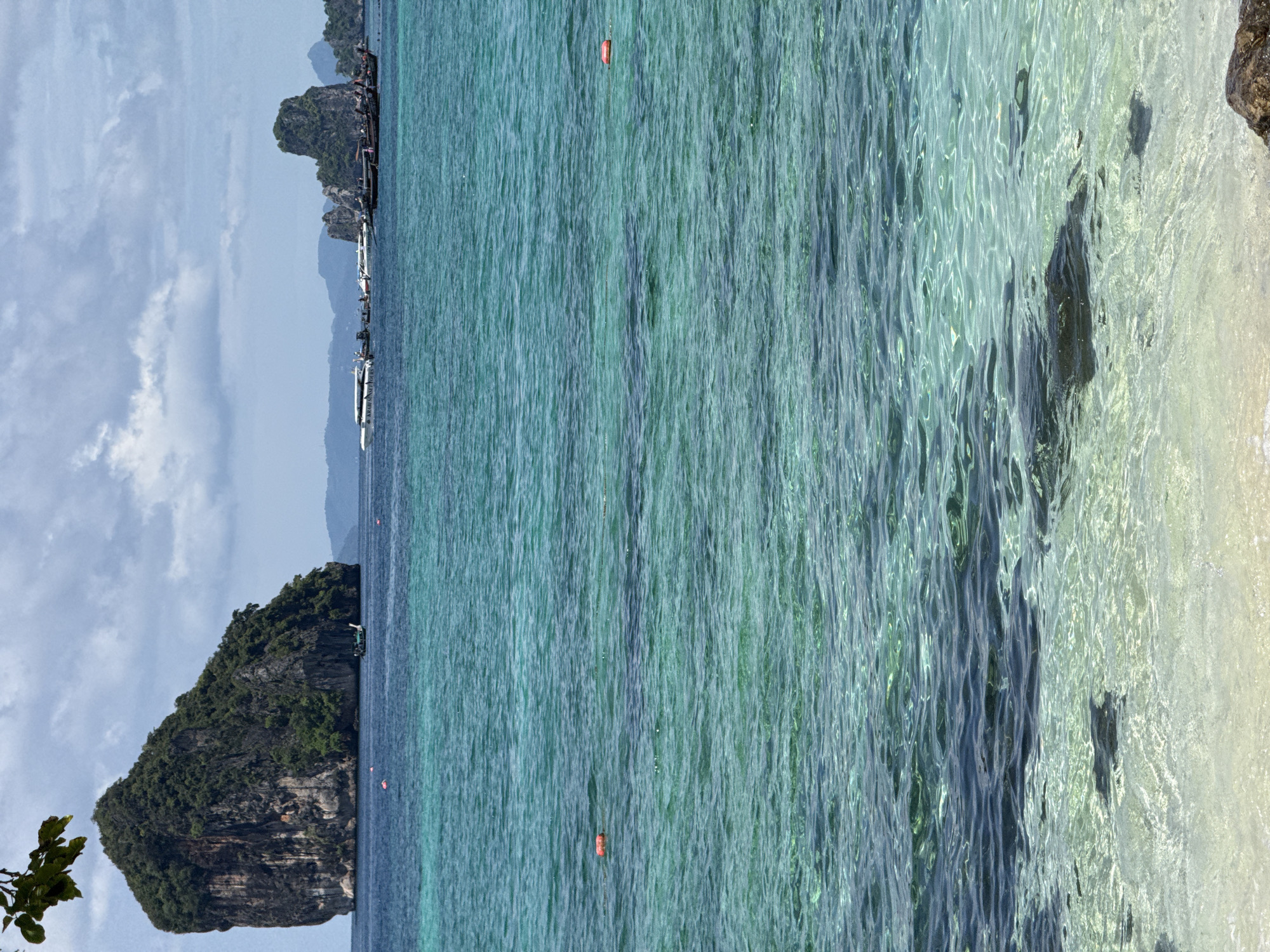 In Ao Nang, Thailand, the image depicts a tranquil ocean scene with several islands in the distance. The foreground is dominated by clear, turquoise water, allowing visibility of the sandy bottom and some rocks. Further out, a cluster of limestone karsts rises from the sea, covered in lush vegetation. Several longtail boats and speedboats are anchored or moving near the islands, suggesting tourist activity. Orange buoys are visible floating on the water's surface. The sky above is partly cloudy, with white clouds against a pale blue backdrop. A tree branch with green leaves is visible in the upper left corner, framing the scene.