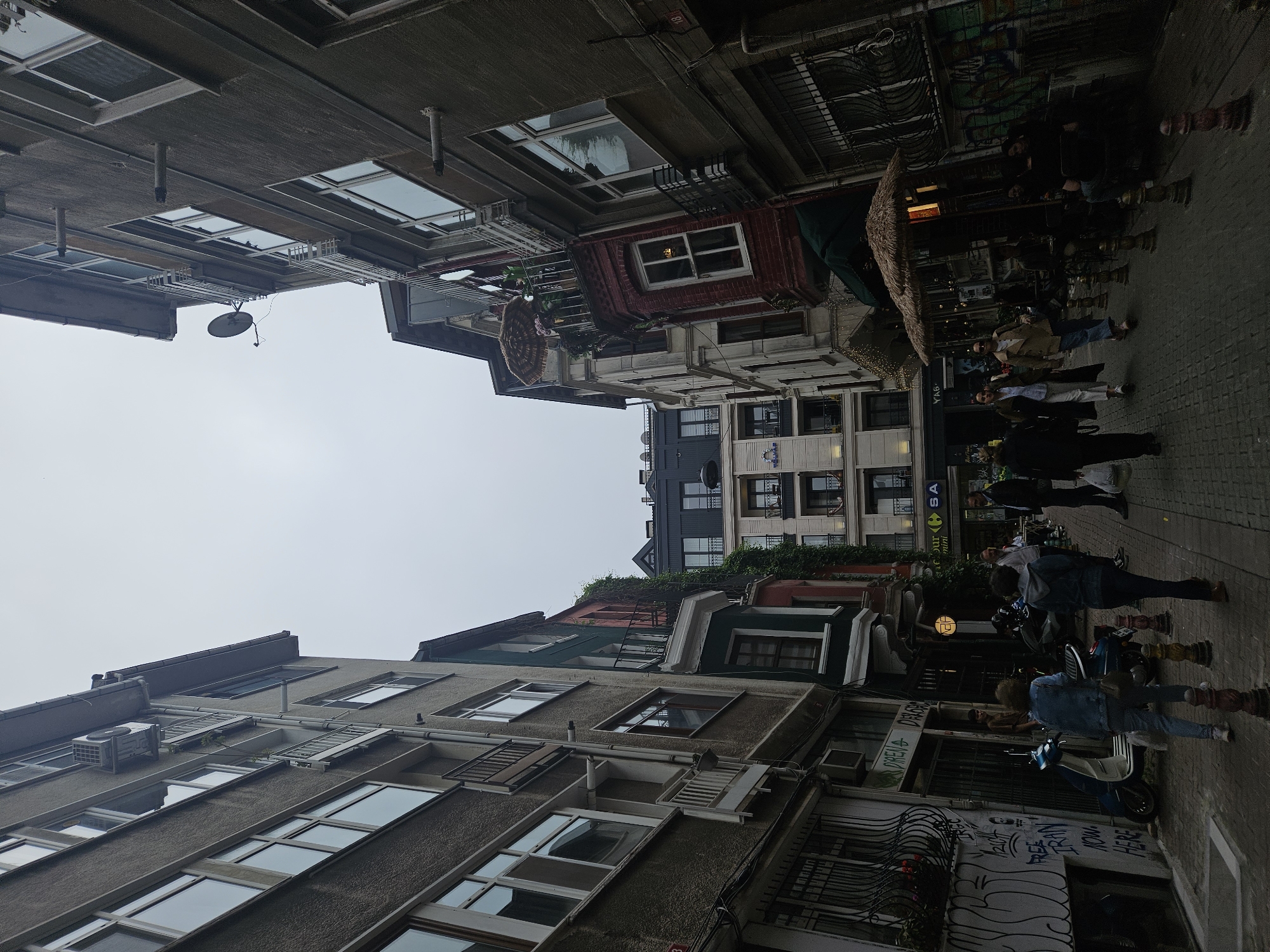 The image depicts a narrow street scene in Kadıköy, Turkey, showcasing a mix of aged buildings and street life. The buildings exhibit a variety of architectural styles and states of repair, with visible windows, balconies, and exterior fixtures. There are people walking along the paved street, and the presence of outdoor seating suggests cafes or restaurants. Some graffiti is visible on the lower parts of the buildings. The sky is overcast.