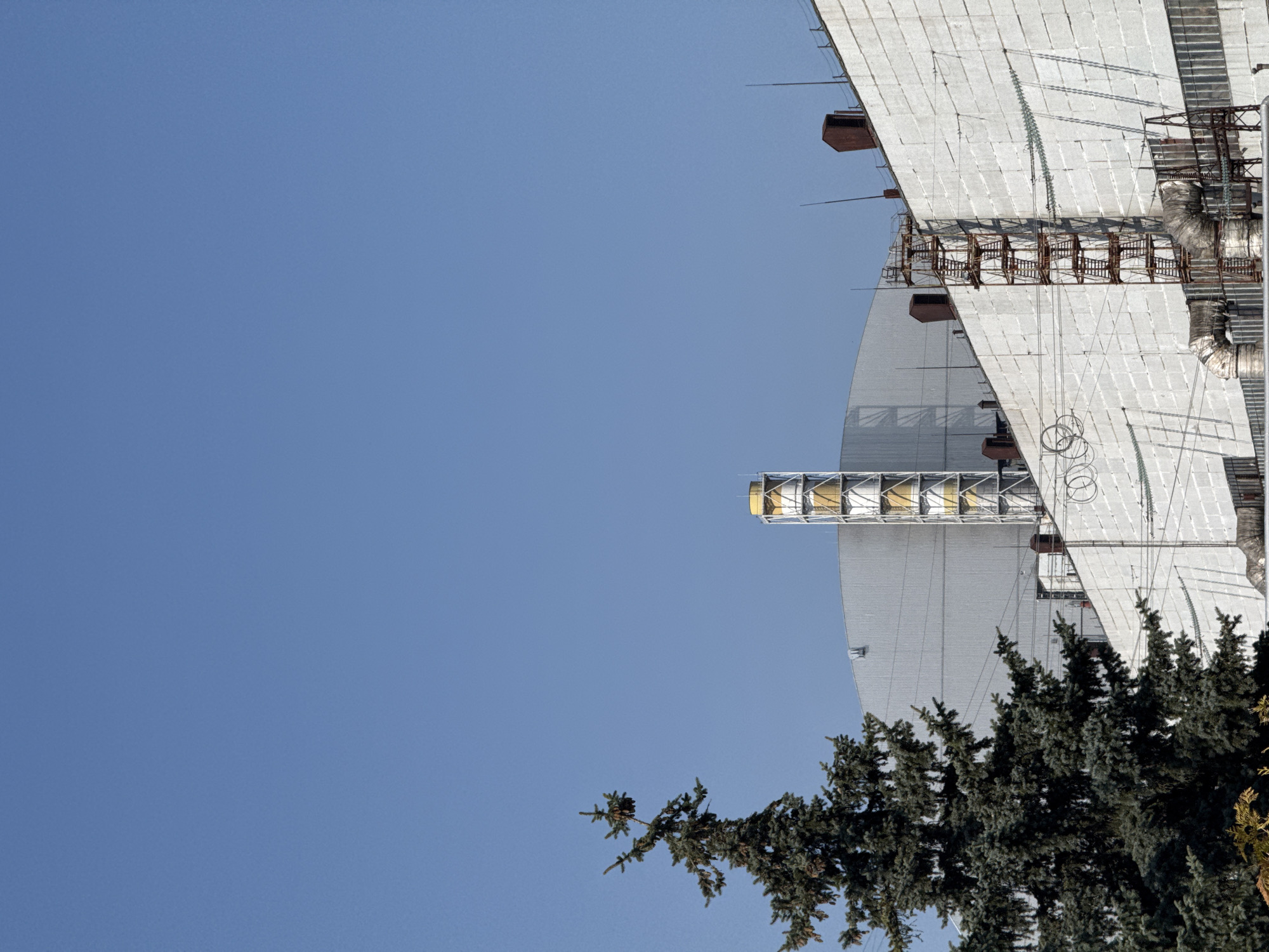 The image shows the Duga radar station in Pryp'yat', Ukraine, viewed from a low angle against a clear blue sky. Part of the massive, grid-like structure dominates the right side of the image, with several levels of metal scaffolding and antenna components. The radar station's surface is composed of numerous rectangular panels. In the bottom right, evergreen trees partially obscure the base of the structure. The orientation of the image is off-kilter, rotated counterclockwise.