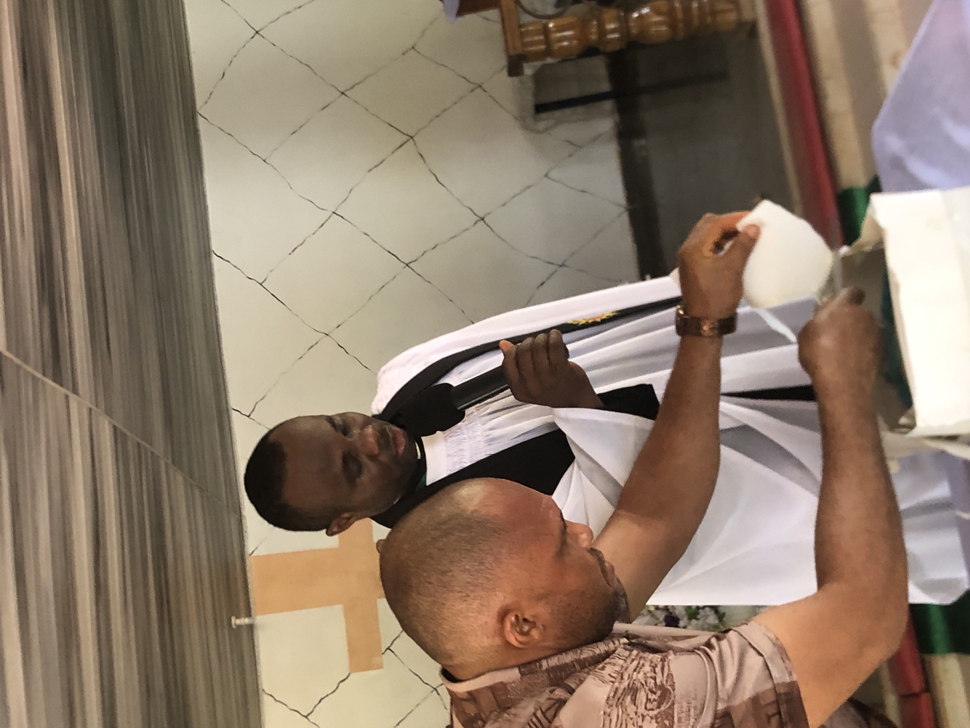 Two men are present. One man is a priest, as indicated by the black robe and white collar. He is holding a microphone. The other man is bald and appears to be pouring something from a white container onto the priest. The environment is a building, possibly a church, with a tiled floor. The image was taken in Ukwu-Abwa, Nigeria.