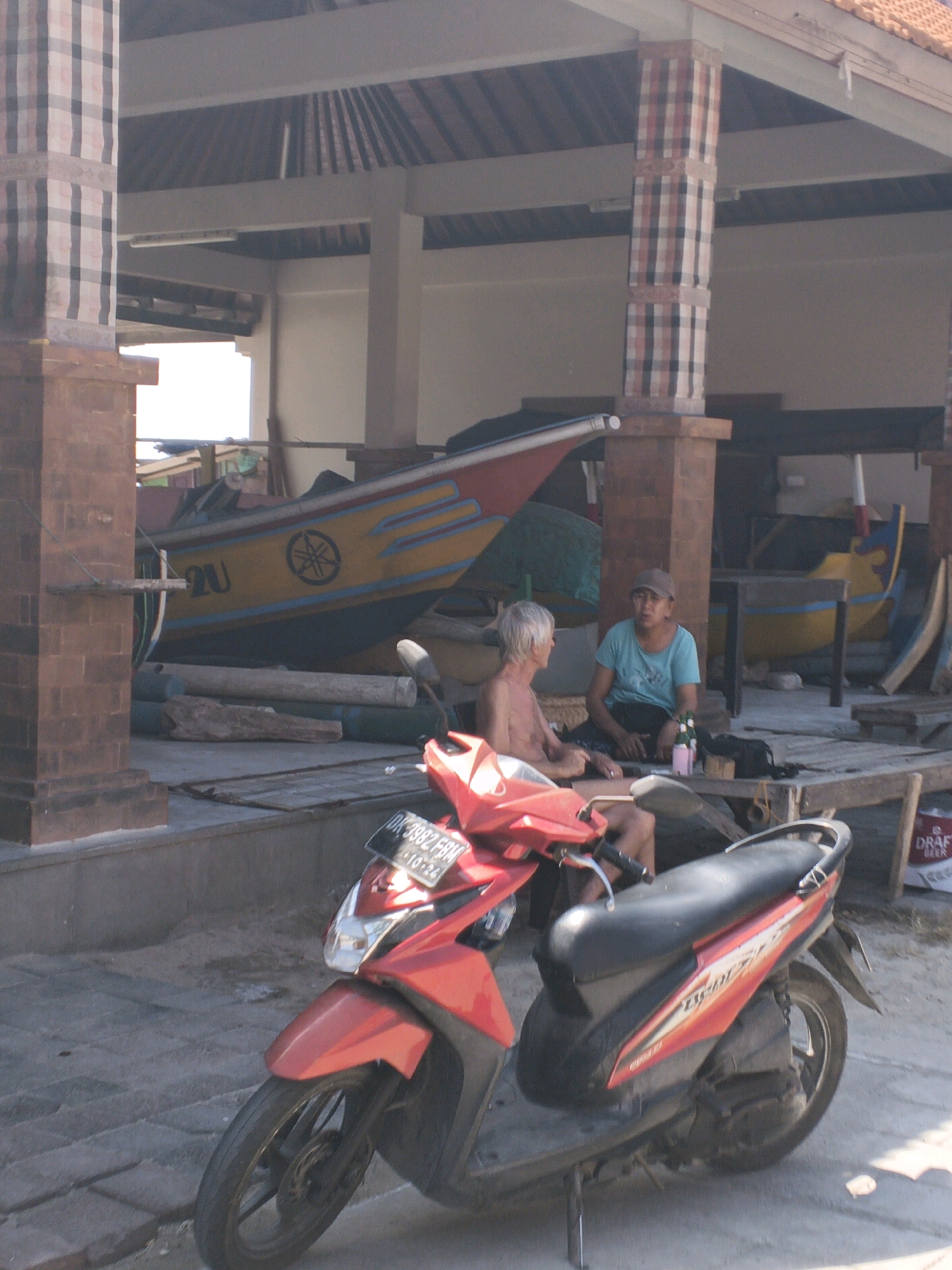 In Kuta Selatan, Indonesia, a red scooter is parked on a paved surface in front of an open-air structure with a tiled roof supported by brick pillars adorned with checkered fabric. Two individuals are seated on a wooden platform within the structure. To the left, a man with short, light-colored hair, wearing no shirt, is seated. To the right, another person, wearing a blue t-shirt and a dark cap, is seated. Between them are a few bottles and other items. Behind the individuals, a yellow and blue boat is partially visible, along with other boats and wooden structures. A white bag with "DRAFT BEER" printed on it is visible in the background.
