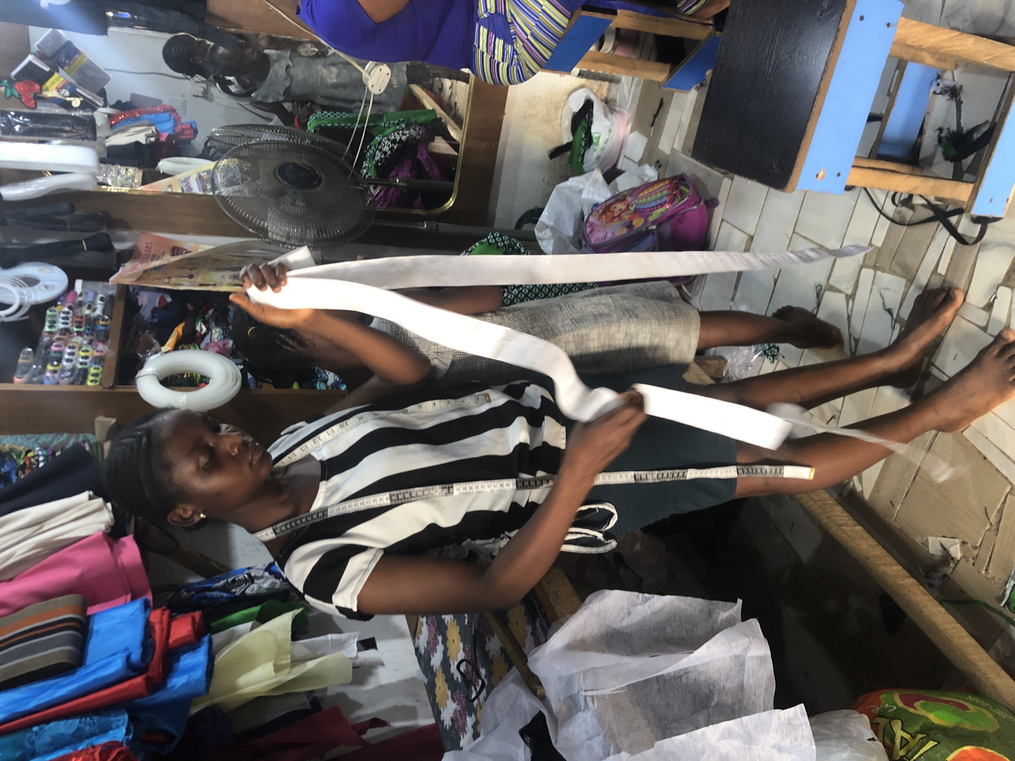 In a cluttered shop in Ado, Nigeria, a young Black woman with braided hair is working. She wears a black and white striped shirt and a tape measure around her neck. She is holding a long, white strip of fabric, seemingly measuring or cutting it. In the background, another young Black girl in a grey dress stands nearby. The shop is filled with various items including bolts of fabric, spools of thread, and sewing supplies, suggesting it is a tailor's shop or fabric store. A fan is visible, and shelves are packed with goods. The floor is tiled, and the overall impression is one of a busy, functional workspace.