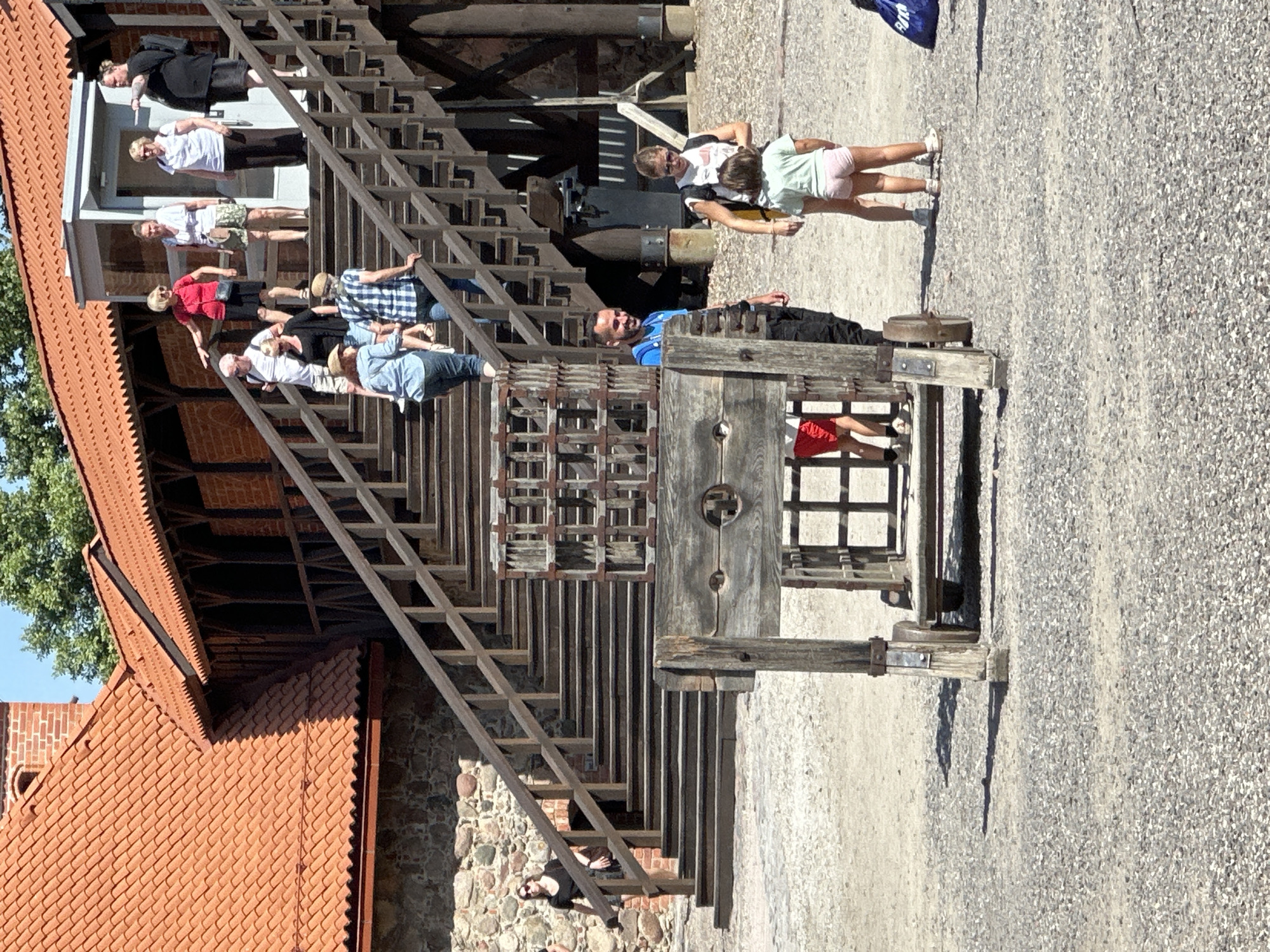 The image depicts an outdoor scene at Trakai Island Castle in Trakai, Lithuania. In the foreground, a wooden pillory, an archaic punishment device, stands on a gravel-covered ground. Behind it, a wooden scaffold structure with metal bars is visible. A man in a blue shirt and black pants stands to the right of the pillory, looking to his left. A woman in a white shirt and shorts with a backpack stands next to a young girl in a light green shirt and pink shorts. Both are looking towards the man. In the background, a wooden staircase with railings leads up to a building with a red tiled roof. Several tourists are visible on the stairs, some ascending and some descending. The environment appears to be a sunny day, with shadows cast on the ground.