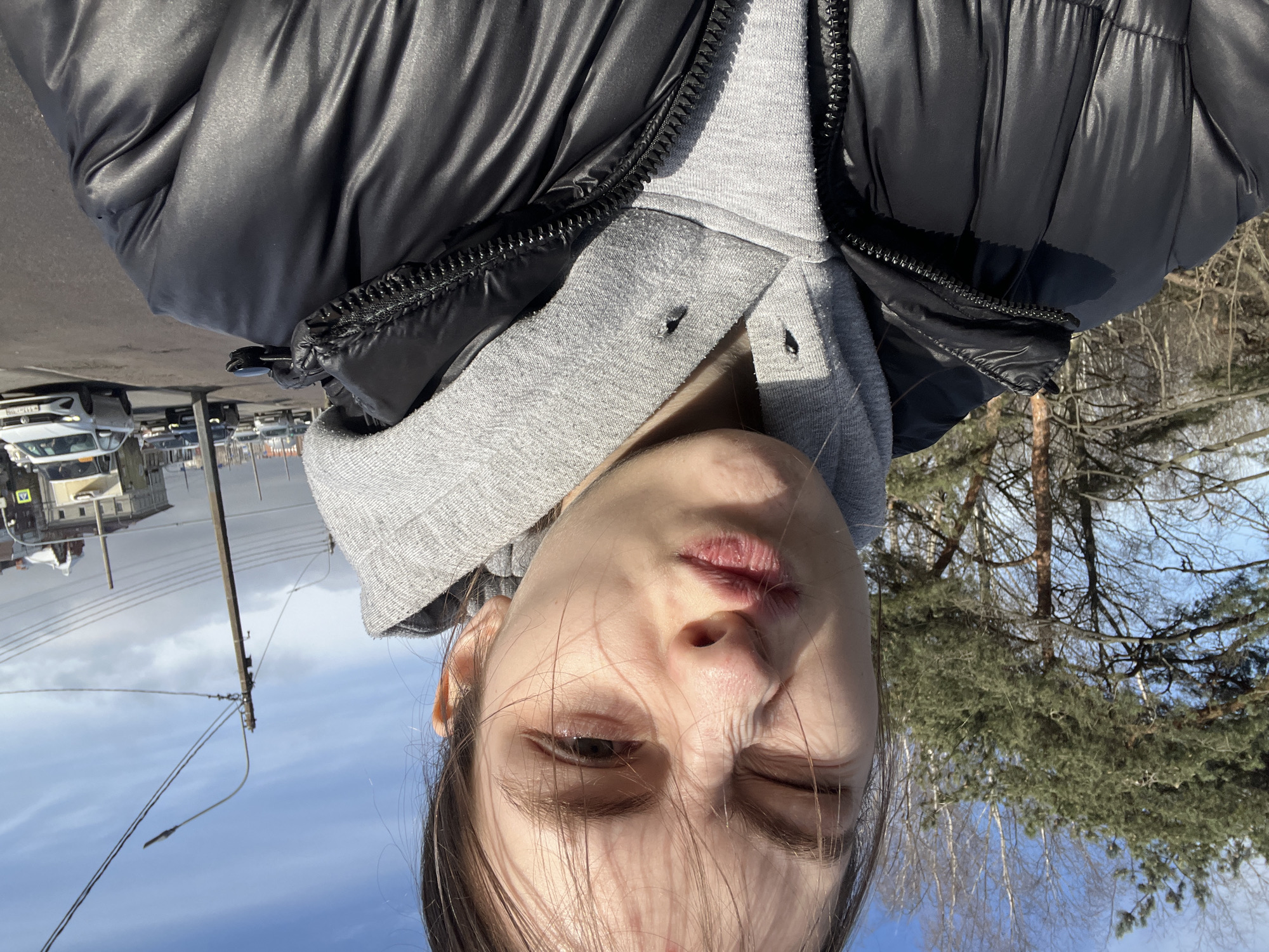 A woman is seen in a selfie-style photograph with her head tilted, wearing a grey hooded sweatshirt under a black puffer jacket. Her face is partially obscured by strands of hair. Behind her, the background is divided into two scenes: one side shows a city scene with buildings, cars, and a utility pole with wires, while the other side displays trees and a blue sky. The presence of trees indicates an outdoor location, potentially near a park or green area. Considering the general appearance of buildings and cars, the image appears to be taken in Saint Petersburg, Russia.