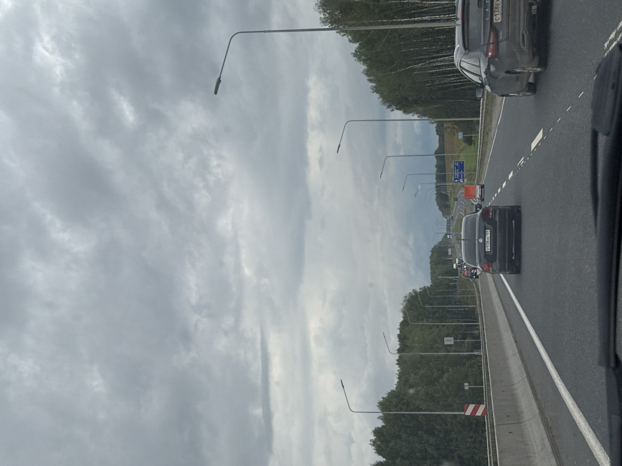 The image depicts a highway scene in Beloborodovo, Russia, under a cloudy sky. The perspective is from inside a vehicle, looking forward. Several cars are visible on the multi-lane road. In the foreground, a black car is in the right lane, and a dark-colored station wagon is in the left lane, slightly ahead. Further down the road, a red truck is in the distance. The highway is bordered by trees on both sides, with a concrete barrier separating the lanes. Streetlights are positioned along the side of the road. Road signs are visible in the distance, including one with blue background and white text. The overall environment suggests a typical highway journey on an overcast day.