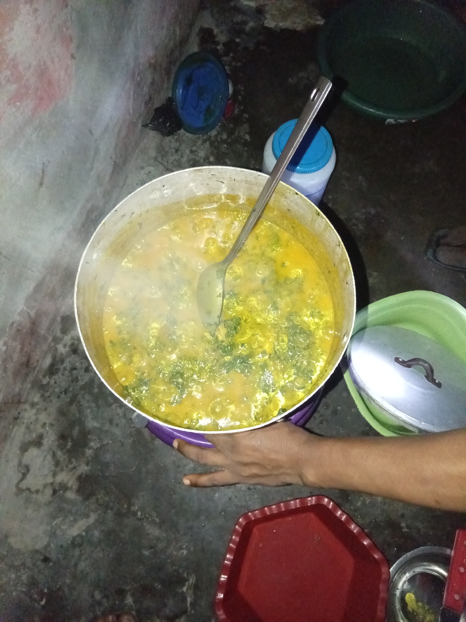 The scene depicts a cooking setup in what appears to be a modest indoor space, likely a kitchen area, in Owerri, Nigeria. A large, silver metal pot filled with a yellow stew containing visible green vegetables is prominently featured. A metal ladle is inserted into the stew. The pot rests on a purple plastic container on a dark grey surface. A hand is seen partially supporting the pot. A green plastic bowl with a lid rests to the right of the pot. Other items in the scene include a red octagonal plastic tray, a small silver dish, and a blue and white container with a lid. The overall lighting is dim, and the background is indistinct.