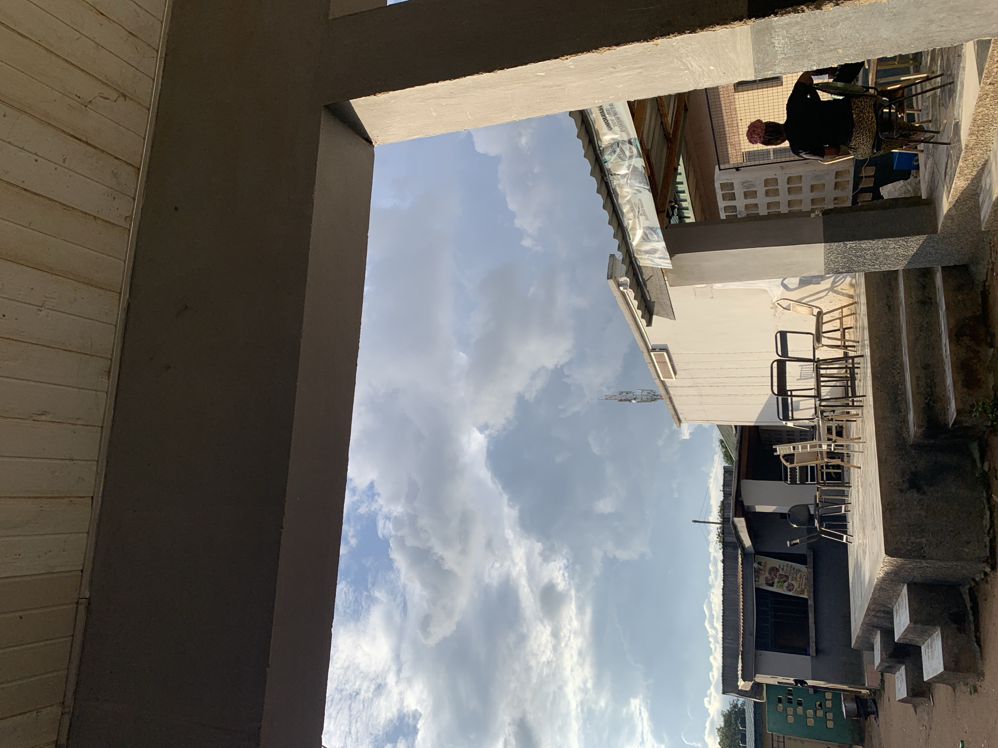 This image, captured in New Stadium, Nigeria, depicts an outdoor scene viewed from beneath a dark grey concrete overhead structure, likely a veranda or canopy, which occupies the upper portion of the frame. A wide concrete pillar frames the right side of the view. The sky beyond is partly cloudy, dominated by large grey and white cumulus clouds with patches of blue visible.
On the left, a single-story building with a dark lower section and lighter upper facade features a corrugated metal roof and a barred window displaying a 'Lady A' poster with food advertisements. In front of this building, a raised concrete platform is accessible via three steps from the left. Several dark-framed chairs are placed on this platform. Further left, a green structure with open blockwork stands near a cooking pot set on a small outdoor burner.
To the right, a white building, also with a corrugated metal roof, supports a communication antenna visible above its roofline. A banner stretched between this building and the foreground concrete structure bears the partially obscured text 'OMO OLUBI YOU ARE DOING WELL'. Beneath the overhead structure on the right, a person with dark skin is seated on a chair, facing away from the camera. They wear a black short-sleeved shirt, a leopard-print or similarly patterned skirt, and a pink or red head covering. The scene is illuminated by natural light, casting soft shadows, consistent with late afternoon conditions.