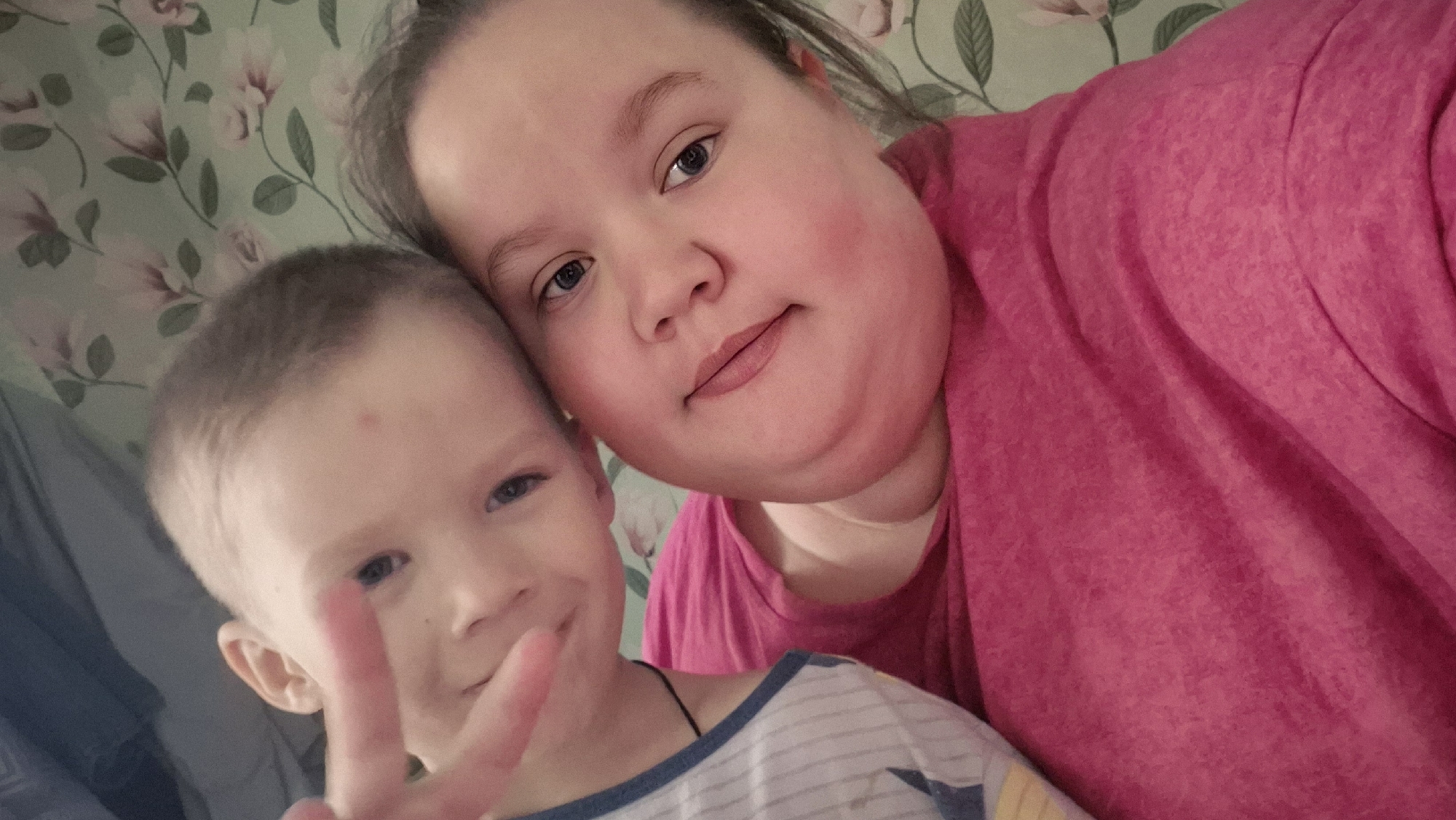 A selfie-style photograph shows two individuals: a child giving a peace sign and an adult. The child, positioned on the left side of the frame, has short light brown hair and blue eyes. The child is wearing a white and blue striped shirt. The adult, to the right of the child and slightly behind, has light brown hair and blue eyes. The adult is wearing a bright pink top. The background features a floral patterned wallpaper with light green and white tones. The location is Большие Кусты, Russia.