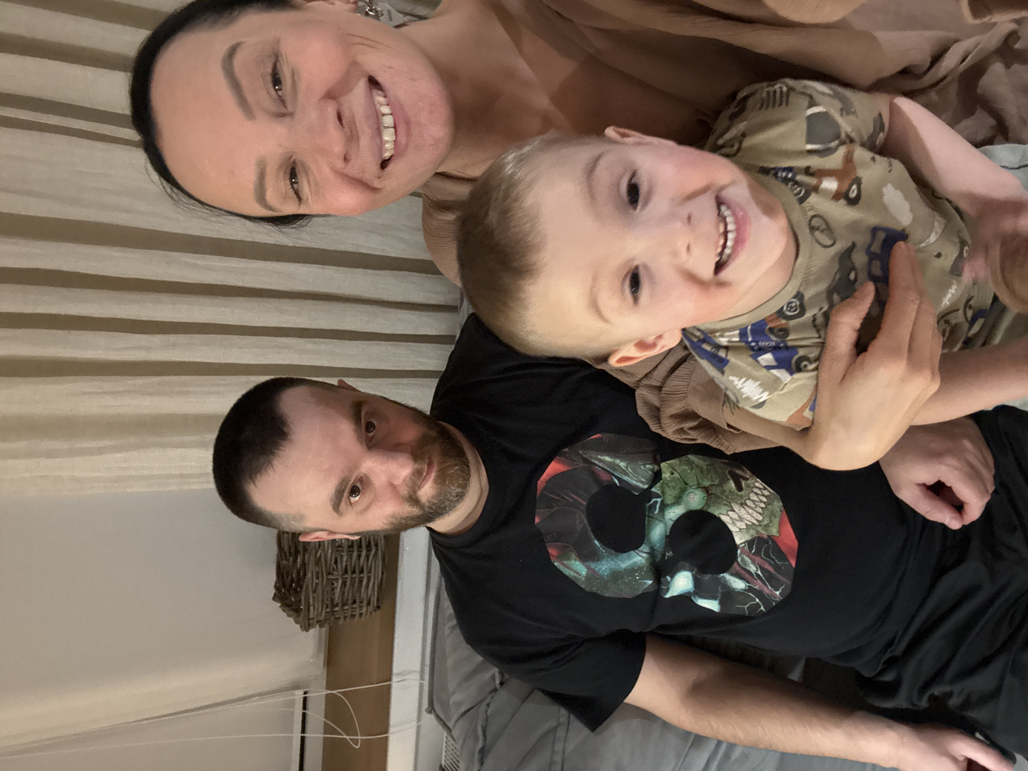 A family selfie in what appears to be an indoor setting. The photo includes three individuals: a woman, a man, and a young child. The woman is positioned at the top of the frame, smiling broadly. The child is in the center, being held. The man is at the bottom, also smiling. He is wearing a black t-shirt. There's a wicker basket visible on the left. The scene appears to be a casual, informal family moment. There is no location information provided. The query for the location in the city of Chaikovsky, Russia is unrelated to the image content, as the location cannot be determined from the image.