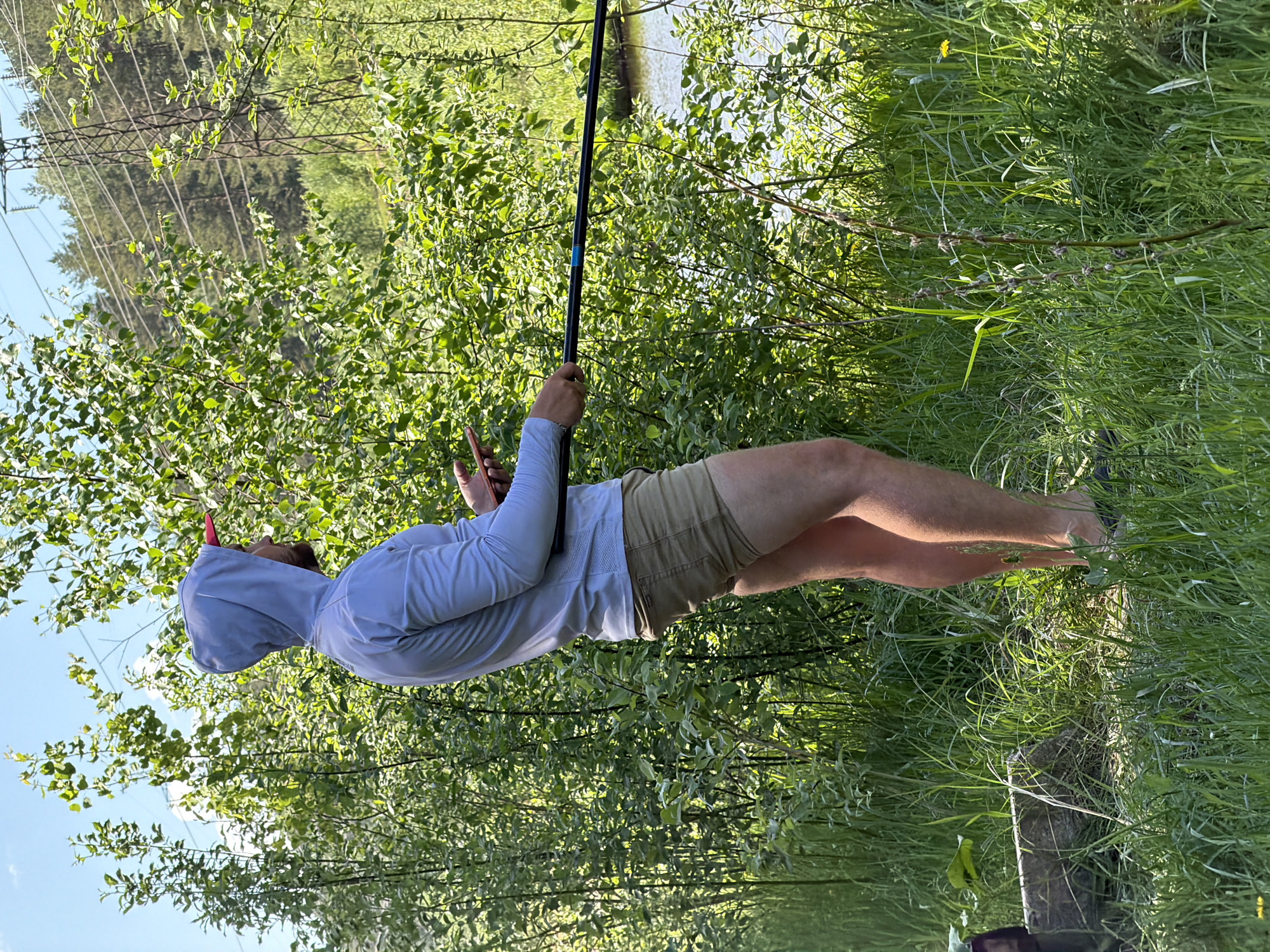 A man stands barefoot in tall grass, holding a long black pole. He wears a light-colored long-sleeved shirt with a hood and khaki shorts. The background features green trees and bushes, with a body of water visible behind the foliage. Overhead, power lines stretch across the sky. The photo appears to be taken in Прикамский, Russia.