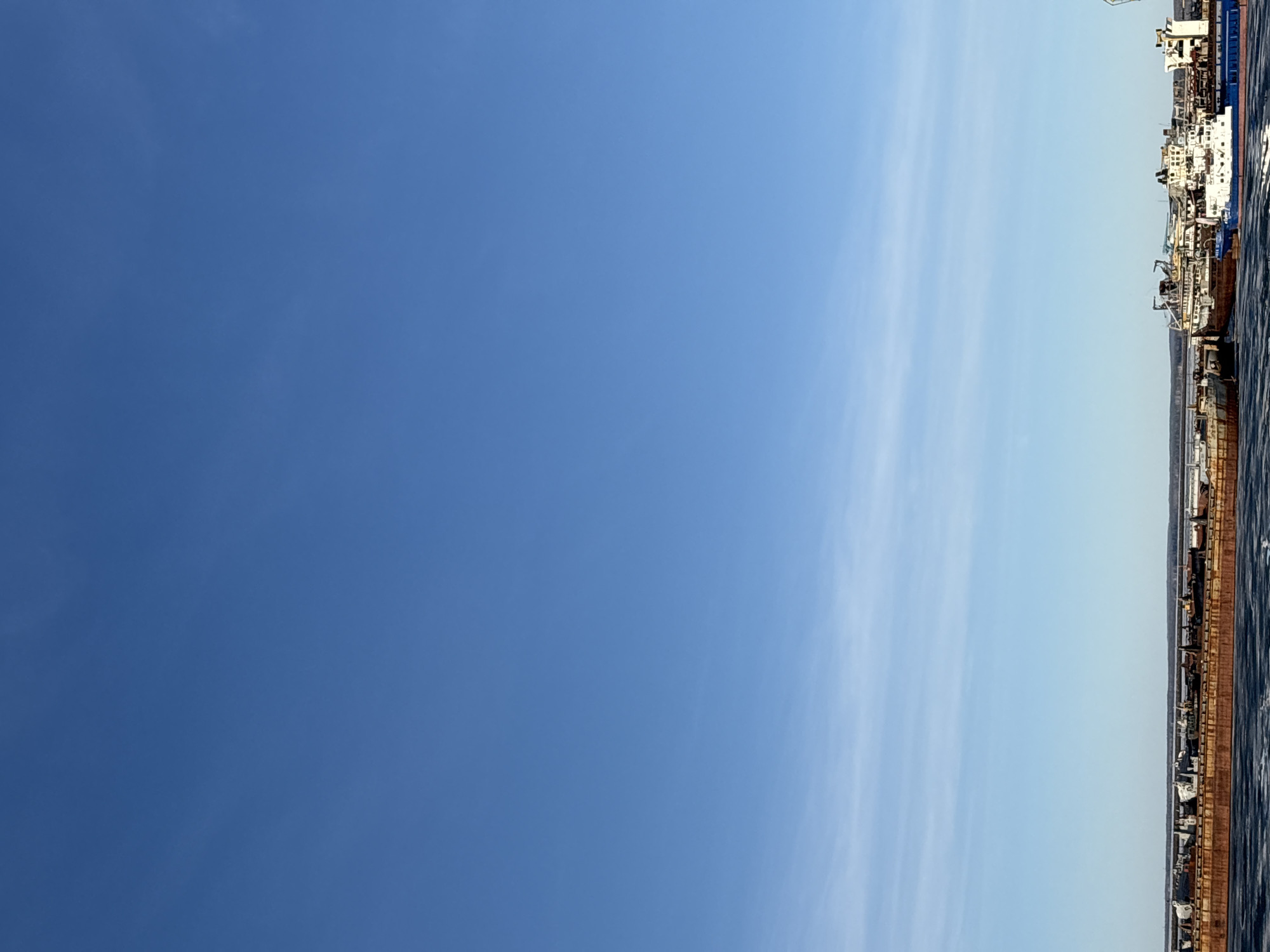 The image shows a waterfront area in the city of Chaikovsky, Russia. The sky, transitioning from a darker blue to a lighter shade, occupies a significant portion of the frame. Along the right edge, there's a dock or shoreline with various structures, possibly ships or industrial facilities, running along it. These structures have a mix of colors, including white, blue, and brown, suggesting a variety of materials and conditions, with visible signs of aging and corrosion. Water or a body of water is visible along the dock. The overall environment appears to be an industrial or port-related setting.