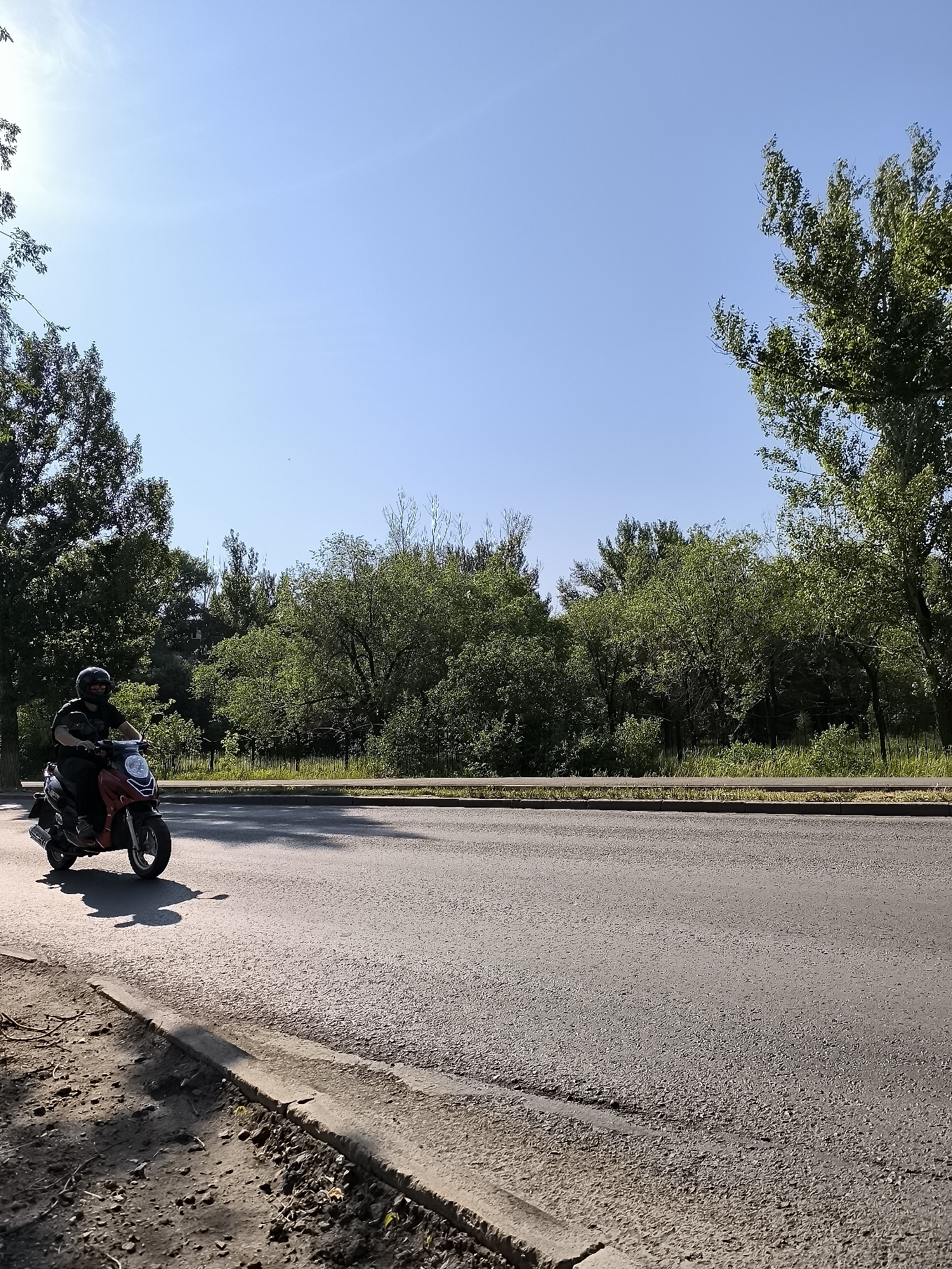 A person is riding a red and black scooter down a paved road on a sunny day. The scooter rider is wearing a black helmet and sunglasses. The road is bordered by a dirt and grass shoulder on the left and a line of trees and bushes on the right. The sky is clear blue. The scene appears to be in an urban or suburban area, possibly in Qarağandı city administration, Kazakhstan.