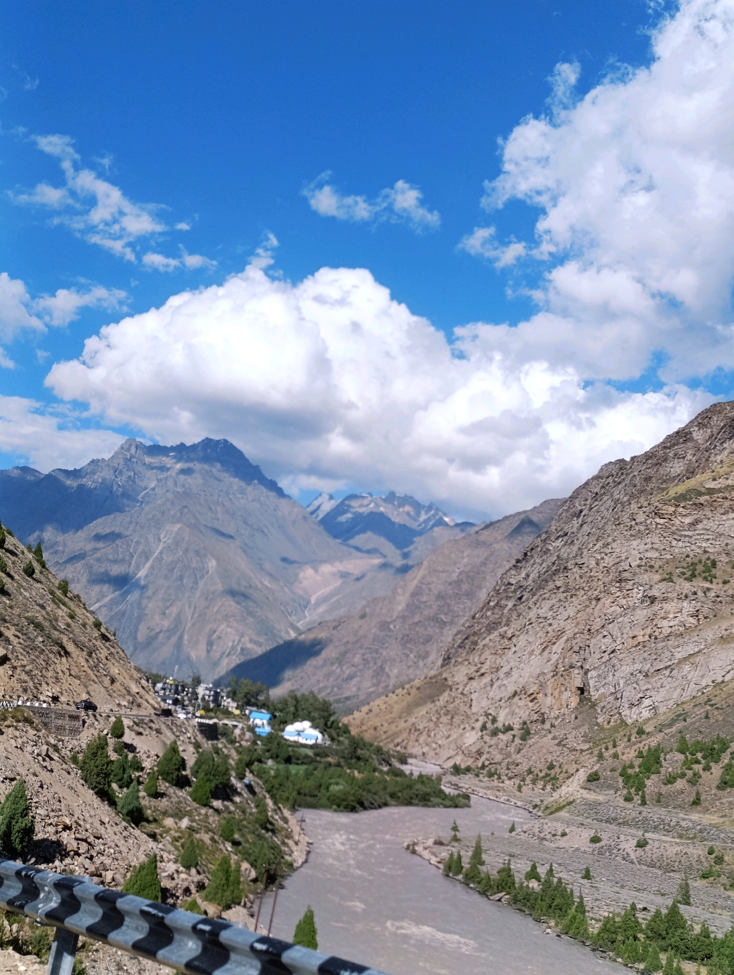 This is a vertical, medium-shot, eye-level photograph of a mountainous landscape with a river and a village. The sky is a vibrant blue with fluffy white clouds. In the foreground, a highway guardrail with black and silver diagonal stripes runs across the bottom of the frame.
The scene depicts a valley with steep, rocky mountains on either side. The left side of the valley is a barren, rocky slope dotted with sparse green vegetation. A winding road is visible on this slope, with several vehicles, including a black SUV, traveling on it. A small village is nestled on the hillside to the right of the road, characterized by white and blue buildings.
The right side of the valley features a more rugged, layered rock face, also with scattered green trees. A wide, murky brown river flows through the valley floor. The river has some small islands with trees and vegetation. In the background, layers of mountains recede into the distance, with some peaks showing signs of snow.
The overall impression is one of a remote, natural setting with human settlement integrated into the landscape. The location is identified as Gemur, India.