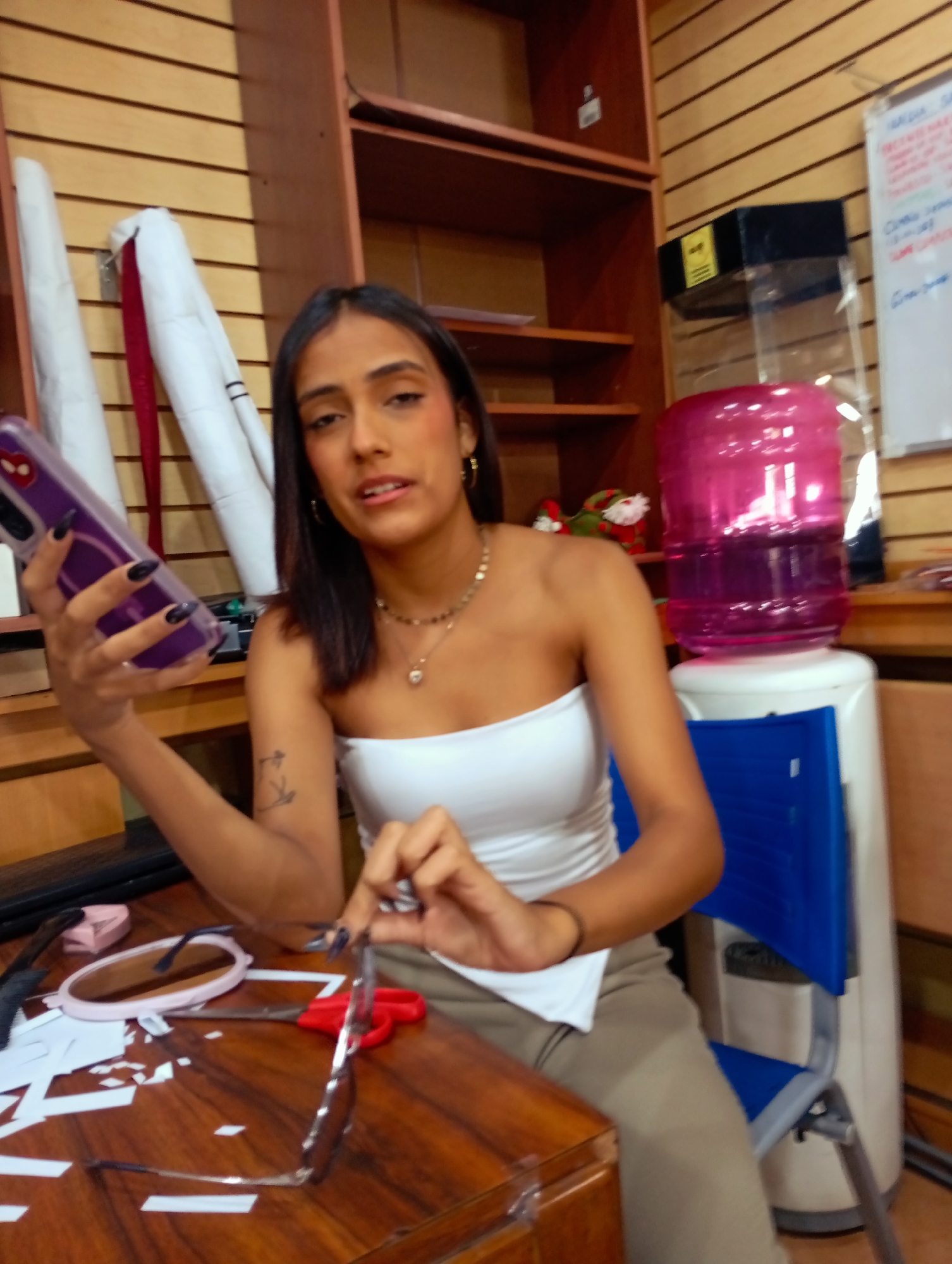 A medium shot depicts a woman seated at a wooden desk indoors, likely in Juan Griego, Venezuela. She holds a purple smartphone in her left hand and scissors in her right, with eyeglasses and paper scraps on the desk in front of her. A white strapless top and light brown pants are visible on her. The desk is cluttered with eyeglasses, a pink case, and paper scraps. Behind her, shelves containing rolled-up white paper and other items can be seen. To her right is a pink water dispenser. The background includes a wall with horizontal wood paneling and a whiteboard.