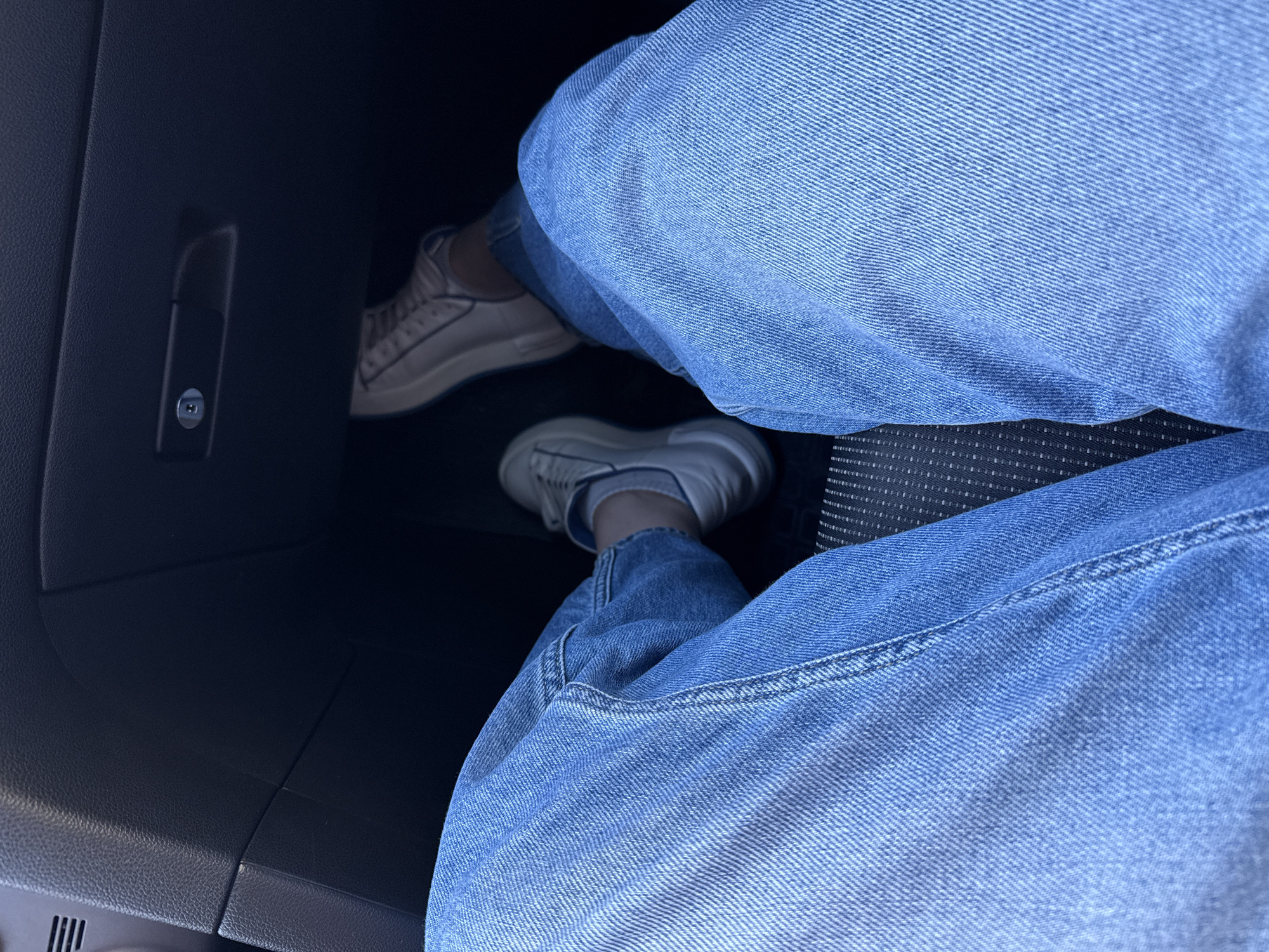 The image shows a person sitting in a car, wearing blue jeans and white sneakers. The person's legs are extended towards the dashboard. The car's interior is dark, with a black glove compartment visible on the left side of the frame. No location information, including Petropavlovsk-Kamchatsky, Russia, is indicated in the image.