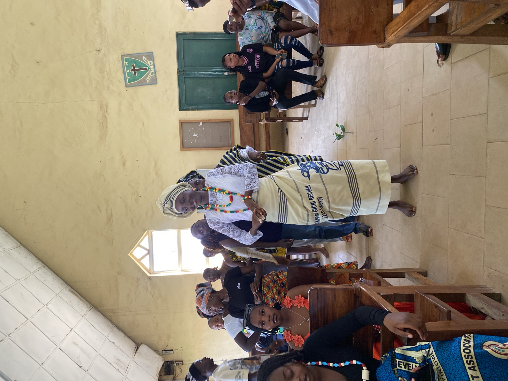 This media file depicts a gathering of people, predominantly women, inside a room that appears to be a church or a community hall. The subjects are seated on wooden benches, some are standing. In the foreground, a woman in traditional attire, including a headscarf and a patterned skirt, is prominently featured. She wears a necklace of colorful beads. Behind her, other individuals are visible, some in casual clothing, others in more traditional outfits. The walls of the room are a pale yellow, and there is a green door and a window with natural light entering the space. A crest with a cross and the words "HOSPITAL VOM" and "N C" is visible on the wall, along with a small plant placed on the floor. Several instances of text are present in the image, including "VICTORY" on a shirt, and "PDA GICHI BECHEH ZADSWORD" on a cloth, and "EVEL VE IRIGW T ASSOCIAT" on a blue patterned fabric. The overall atmosphere suggests a religious service, a celebration, or a community meeting. The time of day is not explicitly indicated, but the natural light suggests daytime. The location cues, such as the crest and the style of the building, point to Dutsen Kato, Nigeria.