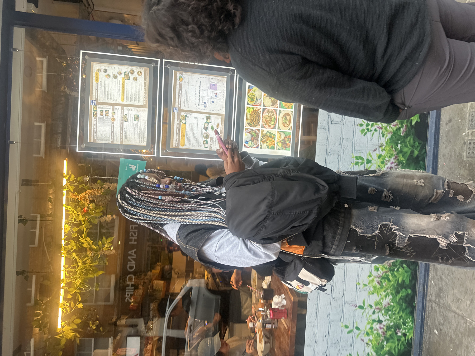 Here is a description of the image:
**Setting:** The image appears to be taken on a street in London, United Kingdom, outside a restaurant or takeaway establishment. The reflection on the windows says "FISH AND CHIPS" in reverse.
**People:** There are at least two people visible. One person is close to the camera, partially obscuring the scene, with dark curly hair and wearing a dark grey jumper and light grey trousers. The second person, facing the window, has elaborate braided hair with colourful beads. This person is wearing a layered outfit consisting of a grey t-shirt, denim jacket, and a dark bomber jacket. They are also wearing ripped jeans.
**Objects:** The main objects in the scene are the menu boards displayed in the windows of the restaurant. The boards feature information about food items, including "THAI Street Noodle", "THAI Street Soup" and a local map. A phone with a pink case is visible in the hands of the second person, appearing to be taking a picture or checking the menu. There's a delivery sign from Deliveroo, indicating that the restaurant offers delivery services.
**Actions and Interactions:** The two people are looking at the menus displayed in the window. The person with the braids is holding a phone.