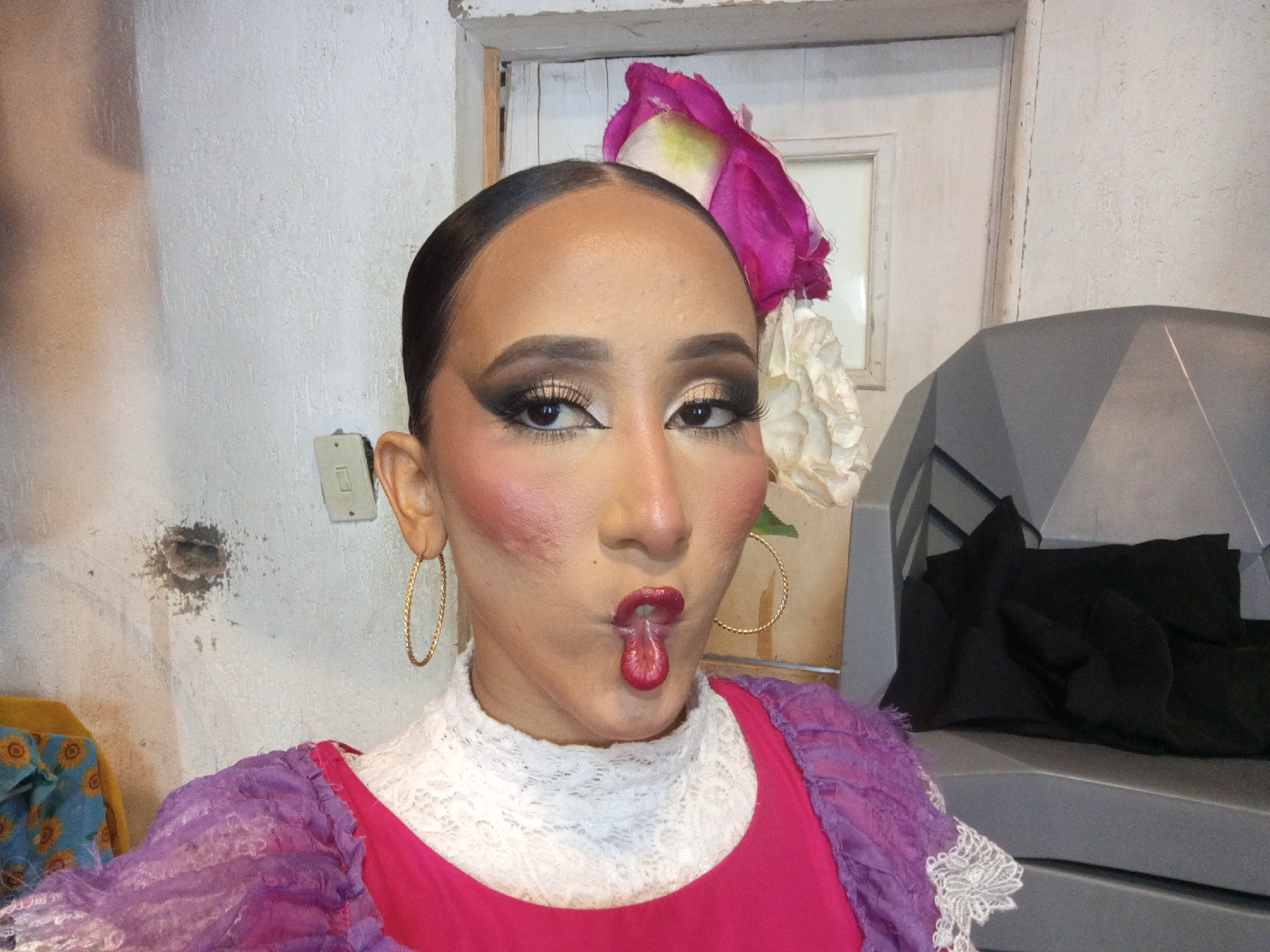 A person, likely a woman, is centered in the frame, taking a selfie. She has dark hair pulled back, heavy makeup, and large hoop earrings. Her attire includes a white lace turtleneck, a bright pink top, and a purple shawl or jacket with lace trim. A large, decorative flower is positioned near her head. She is making a pouting face, sticking her tongue out slightly. The background is a white wall with an electrical outlet and what appears to be a patched hole. A white doorframe with a partially visible window is also visible. To the right, there is a gray, geometric-shaped object with black fabric draped over it. The location is not discernible.