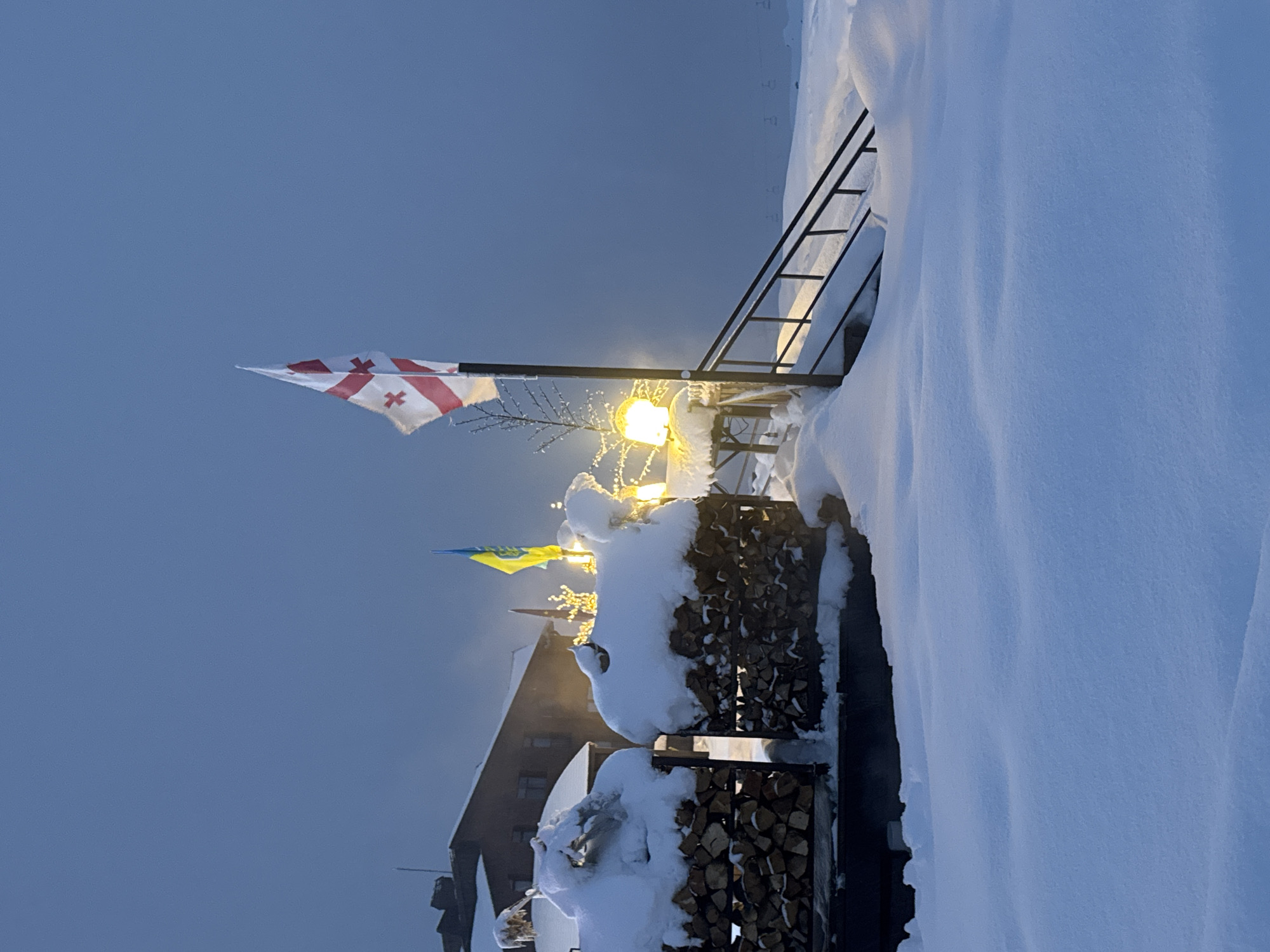 The image depicts a winter scene with a Georgian flag waving in the foreground, alongside a Ukrainian flag partially visible. A glowing light fixture, possibly a street lamp, is situated amidst snow-covered firewood stacks. A building is partially visible in the background, suggesting an urban location experiencing heavy snowfall. The railings and stairs in the scene are also covered in snow. The image does not contain any people or direct interactions. The location is given as Unknown, Unknown.