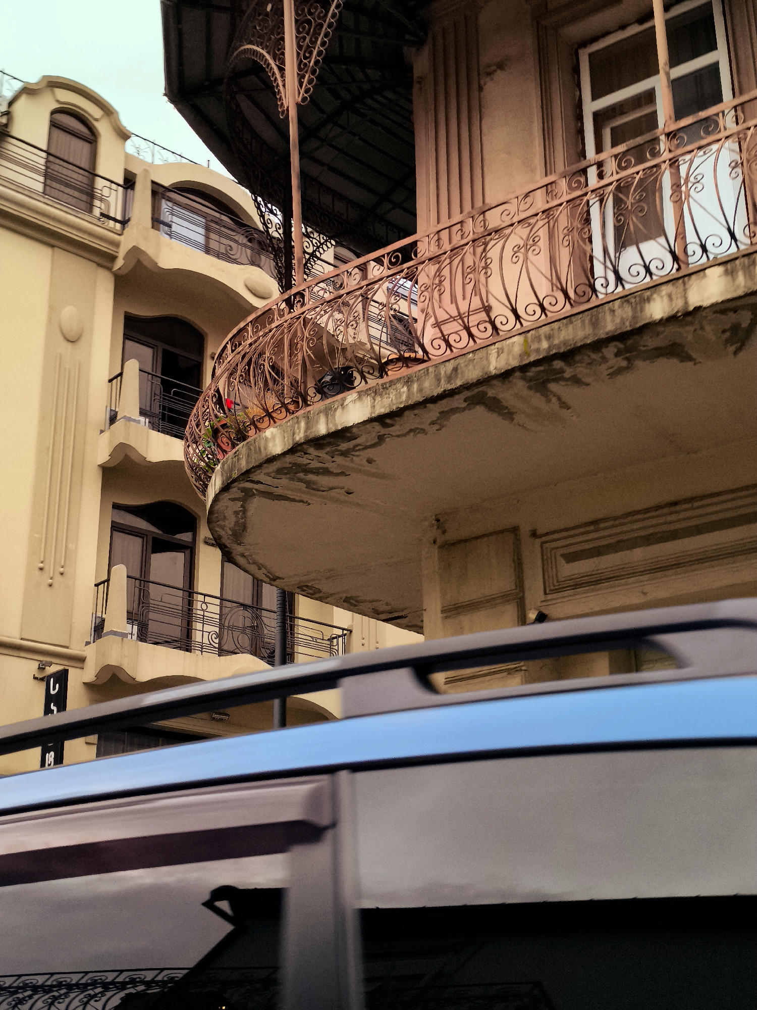 The image shows an ornate, curved balcony with detailed wrought-iron railings extending from a cream-colored building. The balcony's underside is visible, showing signs of wear. Above the balcony, another section of the building features a darker, arched canopy structure. To the left, a multi-story building with arched windows and balconies is visible, painted in a lighter cream color. The lower part of the frame is dominated by the reflective surface of a vehicle, likely a car, with its roof rack clearly visible. The overall scene suggests an urban environment, possibly in Batumi, Georgia, given the architectural style.