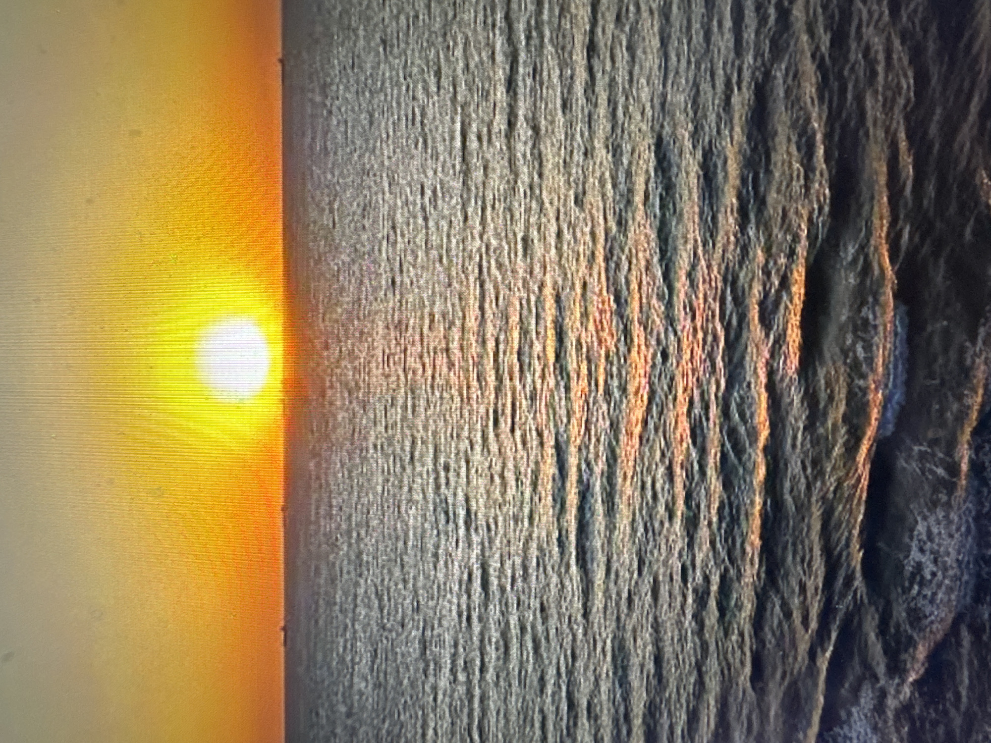 The image depicts a sunset over a body of water. The sun, a bright orb, is positioned low on the horizon, casting a warm, golden glow across the sky and reflecting on the rippling surface of the water. The water itself appears dark and textured with small waves. In the distance, on the horizon, a few faint shapes suggest ships or other vessels. The overall scene is one of natural beauty and tranquility. The location is described as Unknown, Unknown.
