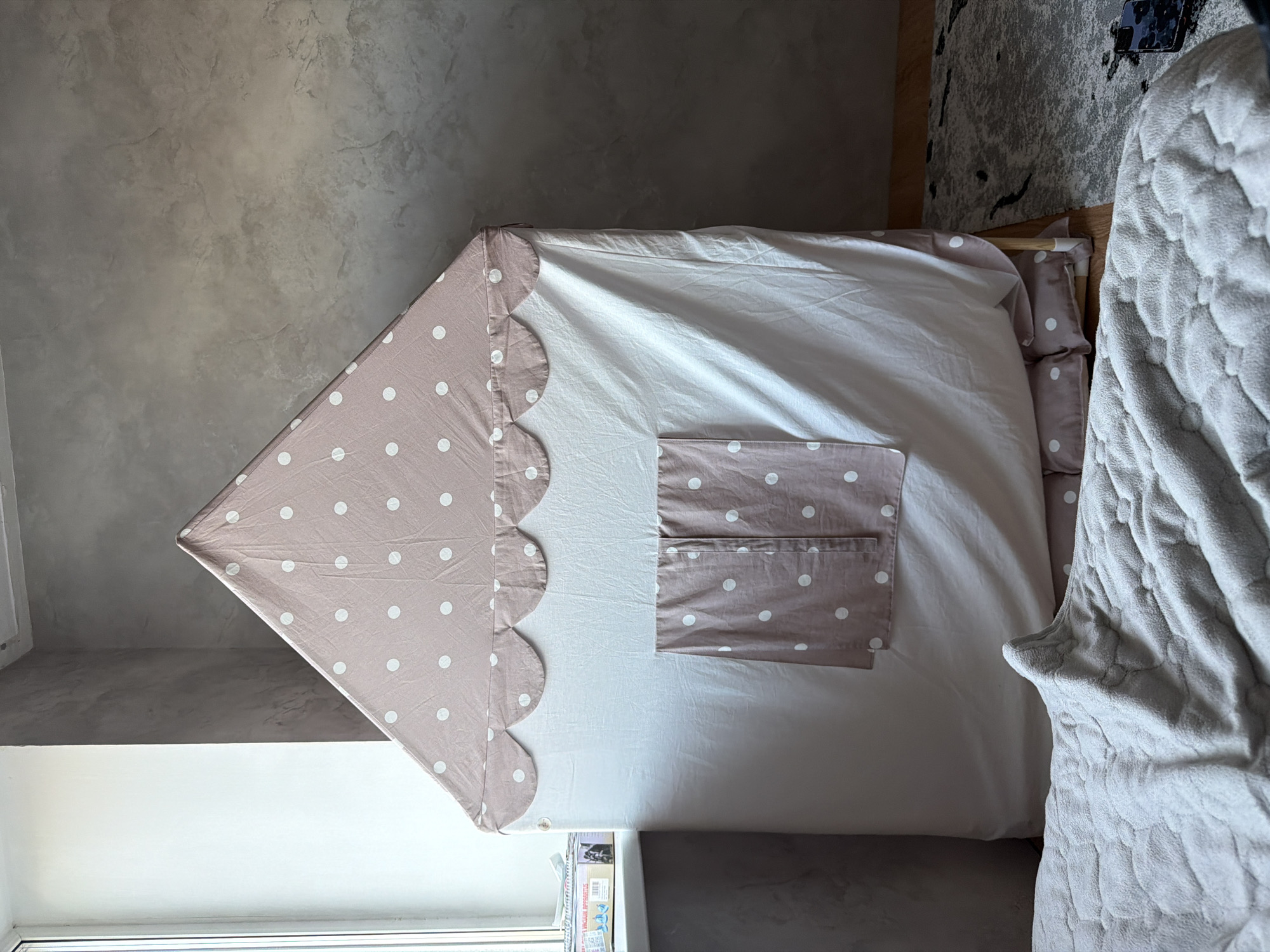 This media file shows a close-up, high-angle view of a children's play tent set up indoors, possibly in a bedroom in Kultaevo, Russia. The tent has a peaked roof shape and is covered with a light-colored fabric featuring a pattern of white polka dots on a taupe background. The front of the tent has a large opening and a decorative scalloped edge along the roofline. There are also two pocket-like details on the front of the tent, also with the polka dot pattern. Inside the tent, white bedding is visible, suggesting it might be used as a bed or a cozy nook.
To the right of the tent, a light gray, textured blanket is draped over something, possibly a bed frame or furniture. The background consists of a textured, concrete-like wall with subtle variations in color and shading. A portion of a dark-colored rug with an abstract pattern is visible in the upper right corner, and a smartphone is placed on it. In the lower portion of the image, a window with a white frame is visible, and below it, some items, possibly books or toys, are stacked. The lighting suggests it is daytime. No people are visible in the frame.