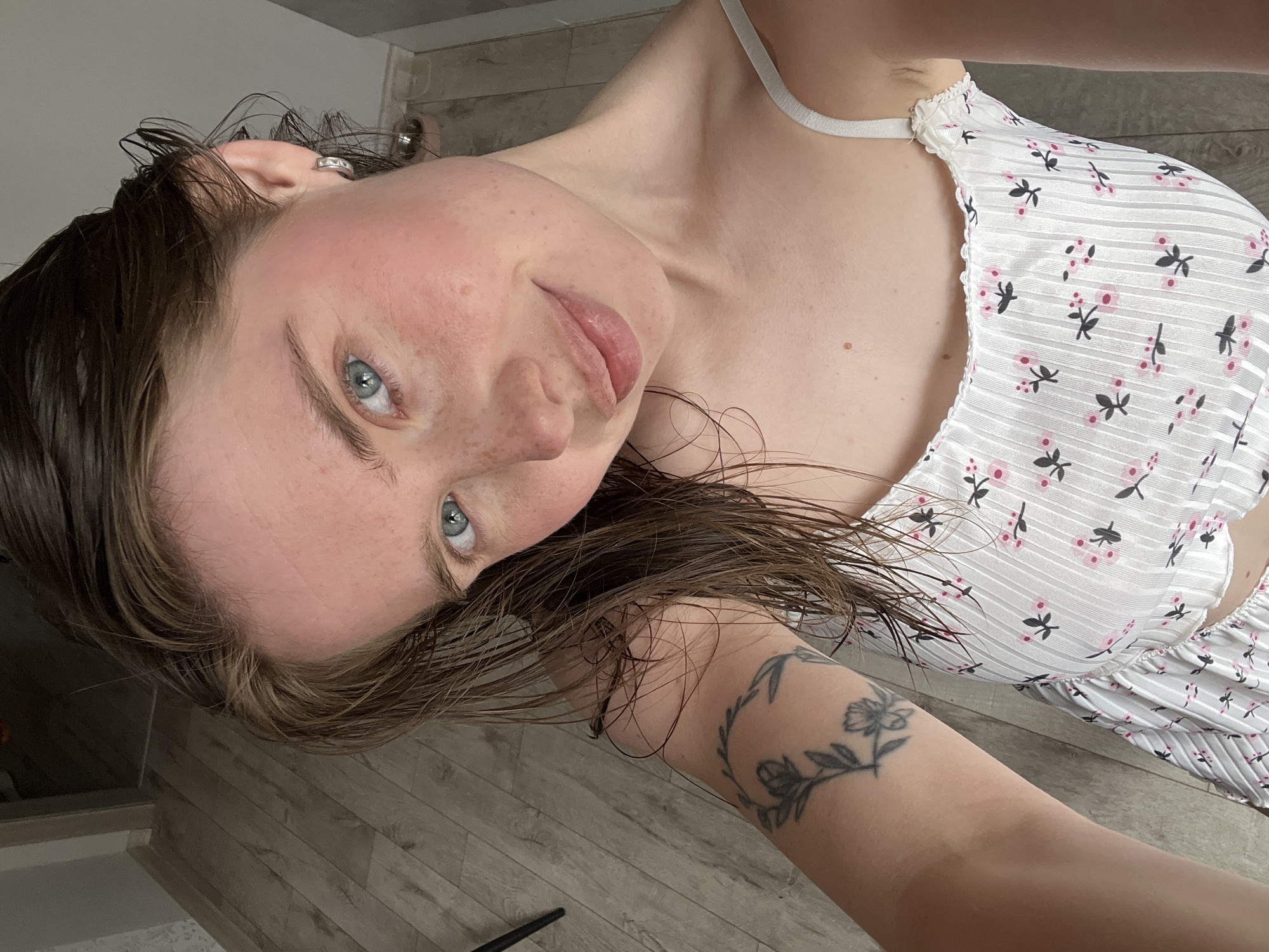 The image is a selfie of a young woman with wet brown hair, blue eyes, and freckles. She is wearing a white floral print dress. She has a tattoo of flowers on her left arm. The background appears to be an indoor setting with wooden flooring. The image was taken in the city of Chaikovsky, Russia.