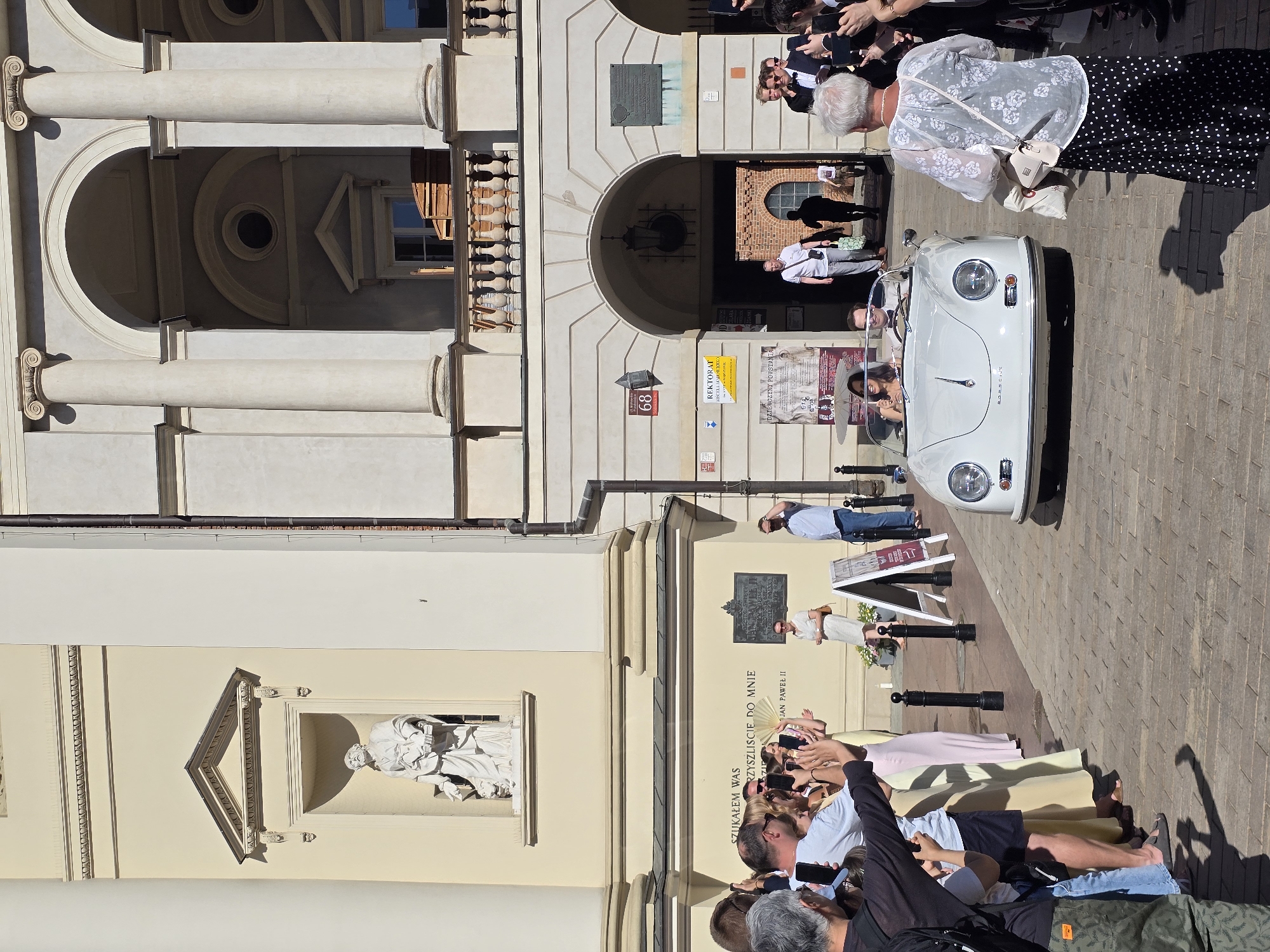 This is a medium shot photograph taken outdoors on a sunny day, depicting a procession passing by a crowd gathered on a cobblestone street in front of a building with classical architecture. The building features large columns, arched entrances, and decorative architectural elements. On the building's facade, there is a statue in a niche and a plaque.
In the foreground, a crowd of people stands along the street, many holding up smartphones to take pictures or videos. They are dressed in casual attire, with some individuals in formal wear, suggesting a special occasion.
The main subject of the procession is a white vintage convertible, possibly a Porsche, carrying a man and a woman who appear to be a couple, dressed in formal attire. The woman is wearing a wedding dress. The car is moving from left to right in the frame.
A woman in a white lace top and a black and white polka dot skirt is standing in the foreground on the right, facing away from the camera and looking towards the car. She is holding a handbag.
In the background, more people are visible walking through an archway into the building, and some figures are seen on a balcony above the arch. The overall scene suggests a wedding or a celebratory event taking place in a historic urban setting in Warsaw, Poland, likely near a significant historical or religious landmark given the architecture. The street is paved with cobblestones, and there are several bollards lining the pathway.
