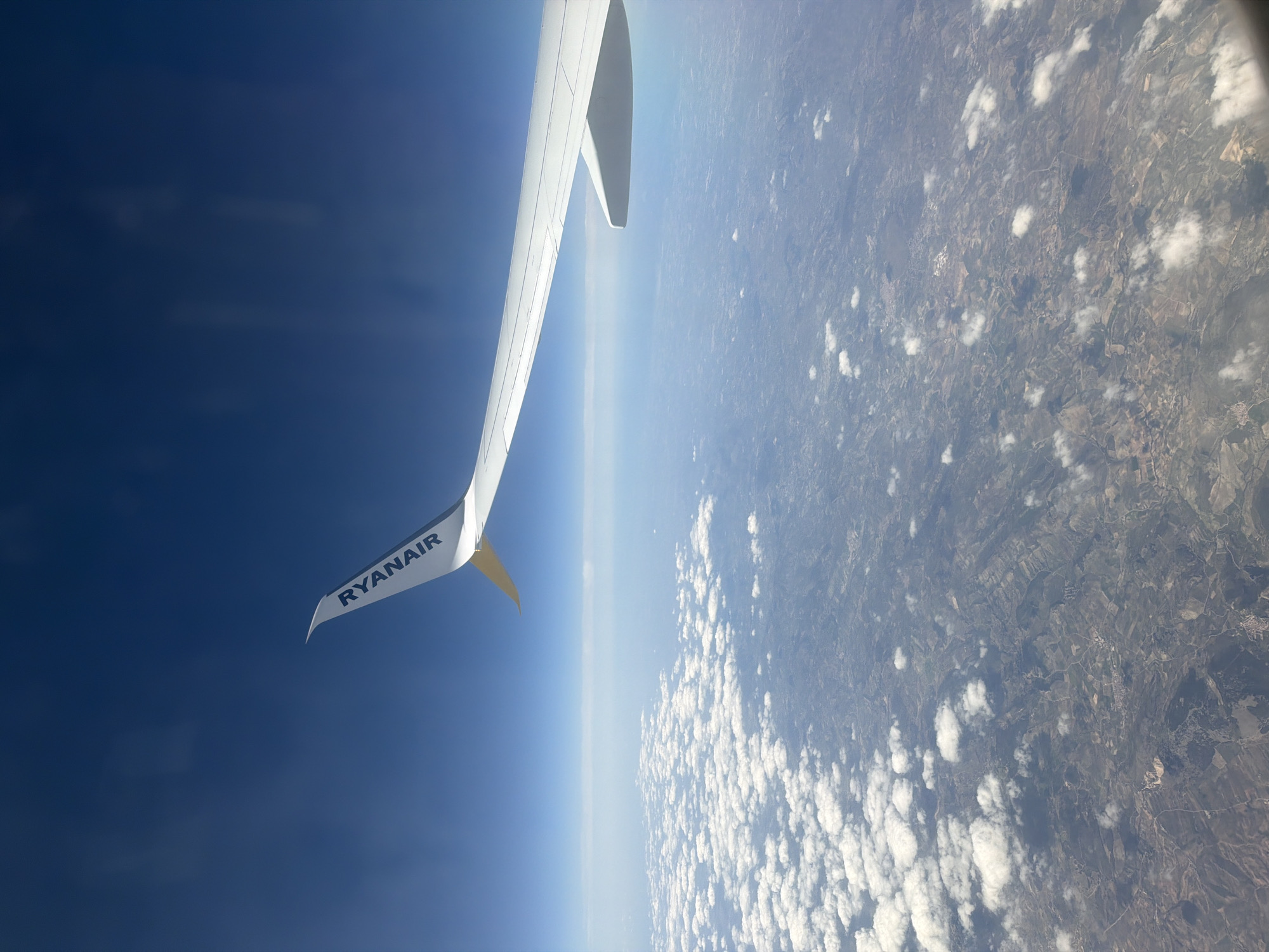 The image presents an aerial view captured from an airplane. A segment of the aircraft's white wing, featuring an upward-curved winglet emblazoned with "RYANAIR" in blue capital letters, is visible in the upper right portion of the frame. The sky above the distant horizon is a clear, deep blue. Below the aircraft's altitude, a scattered formation of white cumulus clouds partially obscures the sprawling landmass. The visible terrain is predominantly rural, characterized by an intricate patchwork of agricultural fields in various shades of green, brown, and tan, interspersed with darker areas of vegetation or small forested sections. Multiple winding roads and several small, low-density human settlements are discernible across the varied landscape. No individuals or specific actions are identifiable on the ground. The aircraft is in flight over this environment. This scene is located over the rural or semi-rural periphery within the administrative boundaries of the municipality of Madrid, Spain, beyond its dense urban core.