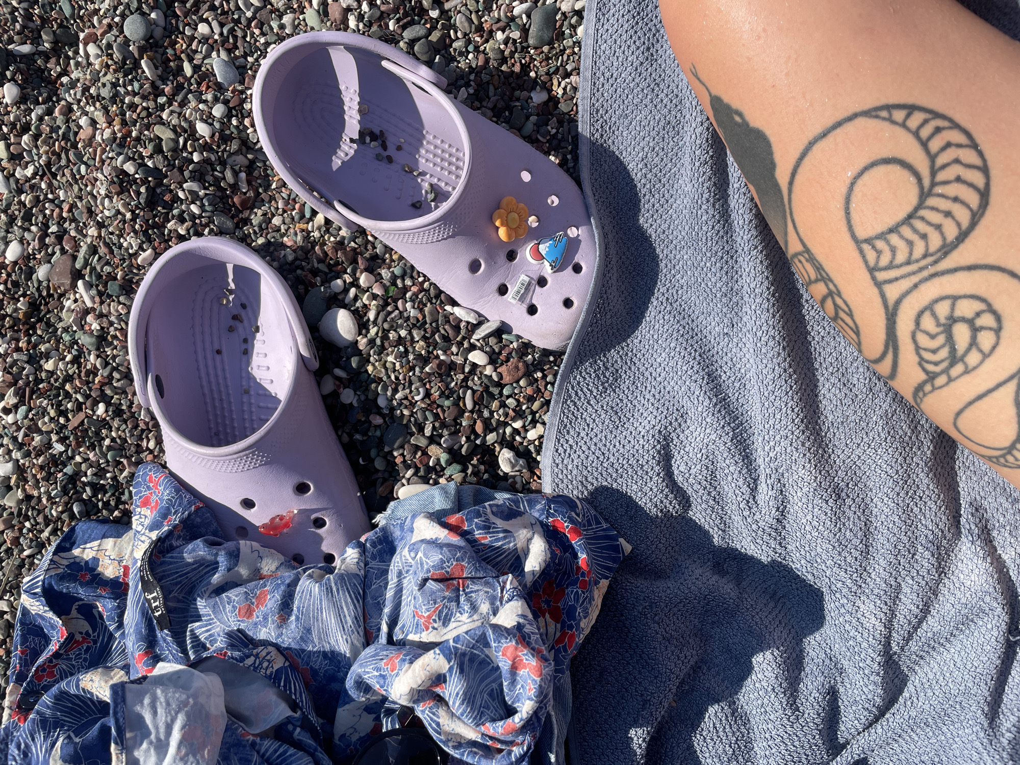 The image shows a pair of light purple Crocs shoes resting on a pebbled beach next to a folded blue and white patterned shirt. A textured blue towel is draped over the pebbles, and a person's arm with a black snake tattoo is visible on the towel. The scene suggests a relaxed day at the beach.
The Crocs have two charms attached: a small orange flower and a blue and white charm with a red detail, possibly a rocket or airplane. The shoes appear to be new, with a white sticker on the side of one shoe. The patterned shirt has a black tag with white text that is partially visible, reading "JEF".
The sunlight casting shadows on the towel and the bright illumination of the pebbles suggest it is daytime, likely sunny. The location cues, such as the pebbled beach and the clothing items, are consistent with a beach setting. The overall mood is one of leisure and enjoyment of outdoor activities.
