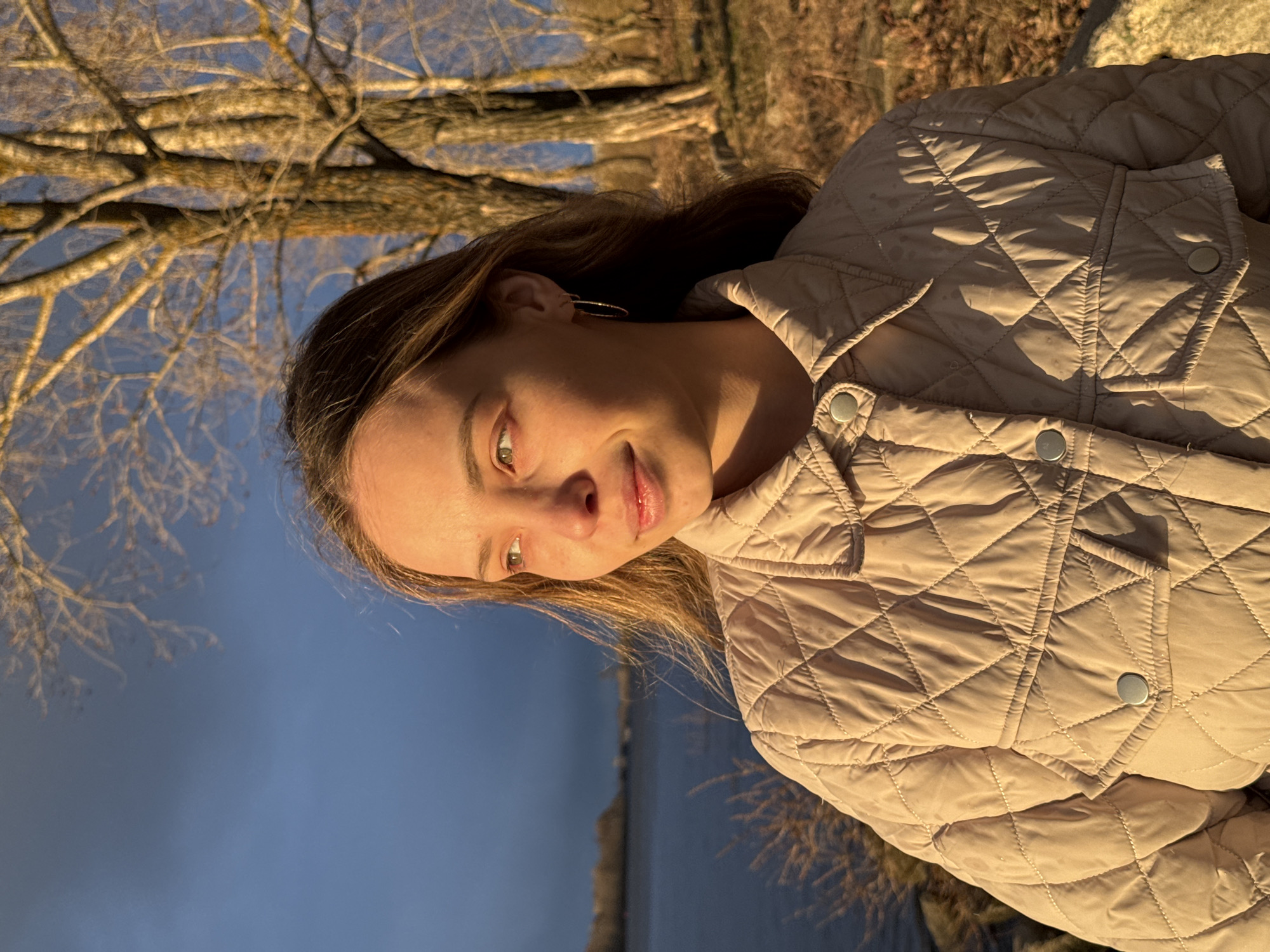 A young woman is captured in a vertical, outdoor portrait during the golden hour, likely in Chaikovsky, Russia. She occupies the right side of the frame, seen from the chest up with her head slightly tilted. Her light brown hair frames a face with fair skin, illuminated by the warm, directional light of the low sun, which casts gentle shadows and highlights her features, including her green or hazel eyes. She has a soft, slight smile and is looking gently away from the camera. She wears a light beige or tan quilted jacket with visible silver snap buttons, suggesting a cool climate. A small hoop earring is visible in her right ear.
The background on the left features a tranquil body of dark blue water, likely a river or lake, stretching into the distance. Above the water, a network of bare tree branches, devoid of leaves, extends across the upper part of the scene against a deep blue sky. This detail, along with the woman's jacket, suggests a season of late autumn, winter, or early spring. The strong, warm light indicates late afternoon or early evening. The scene conveys a peaceful, natural setting. There is no visible text in the image.