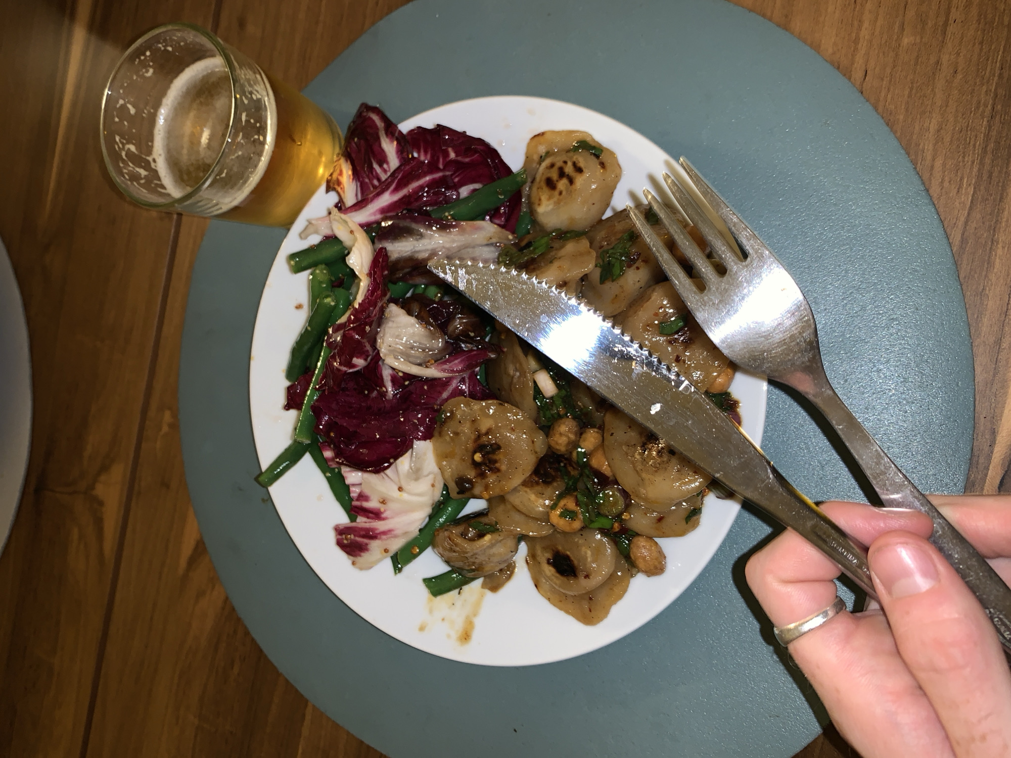 A plate of food is centered in the image, with a glass of beer to the upper left. The plate contains a mix of salad greens, possibly radicchio, green beans, and pan-fried dumplings with herbs and peanuts. A fork and knife are positioned on top of the food, held by a hand. The scene takes place on a round, blue-grey placemat set on a wooden table. No clues suggest the specific city or Argentina. The setting appears to be an informal dining environment.