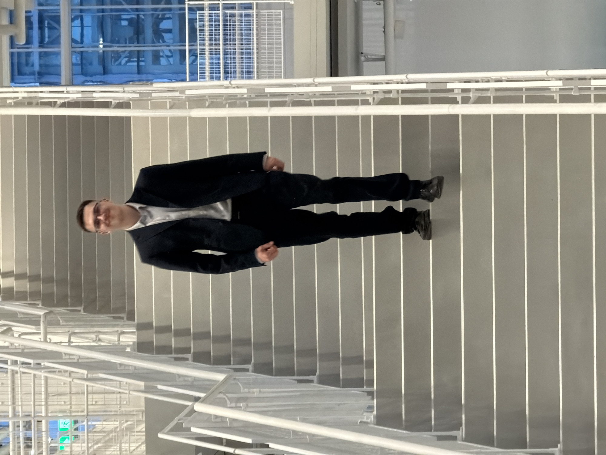 A man dressed in a dark suit and light-colored shirt is standing on a set of stairs. The stairs have a white railing and appear to be indoors. The location is not discernible from the image.