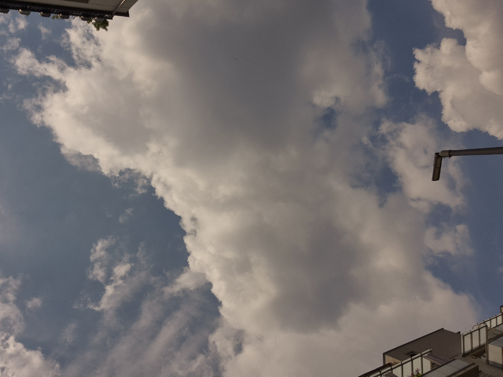 The image captures a view of a cloudy sky with buildings and a streetlamp. In the lower left quadrant, a portion of a modern building with balconies is visible. To the right of the building, a tall, gray streetlamp stands prominently in the lower center of the frame. The sky is filled with a mix of white and gray clouds, with patches of blue sky showing through. A small, dark bird is visible in flight in the upper right quadrant of the sky. The scene appears to be an urban environment, likely during the daytime, given the illumination.