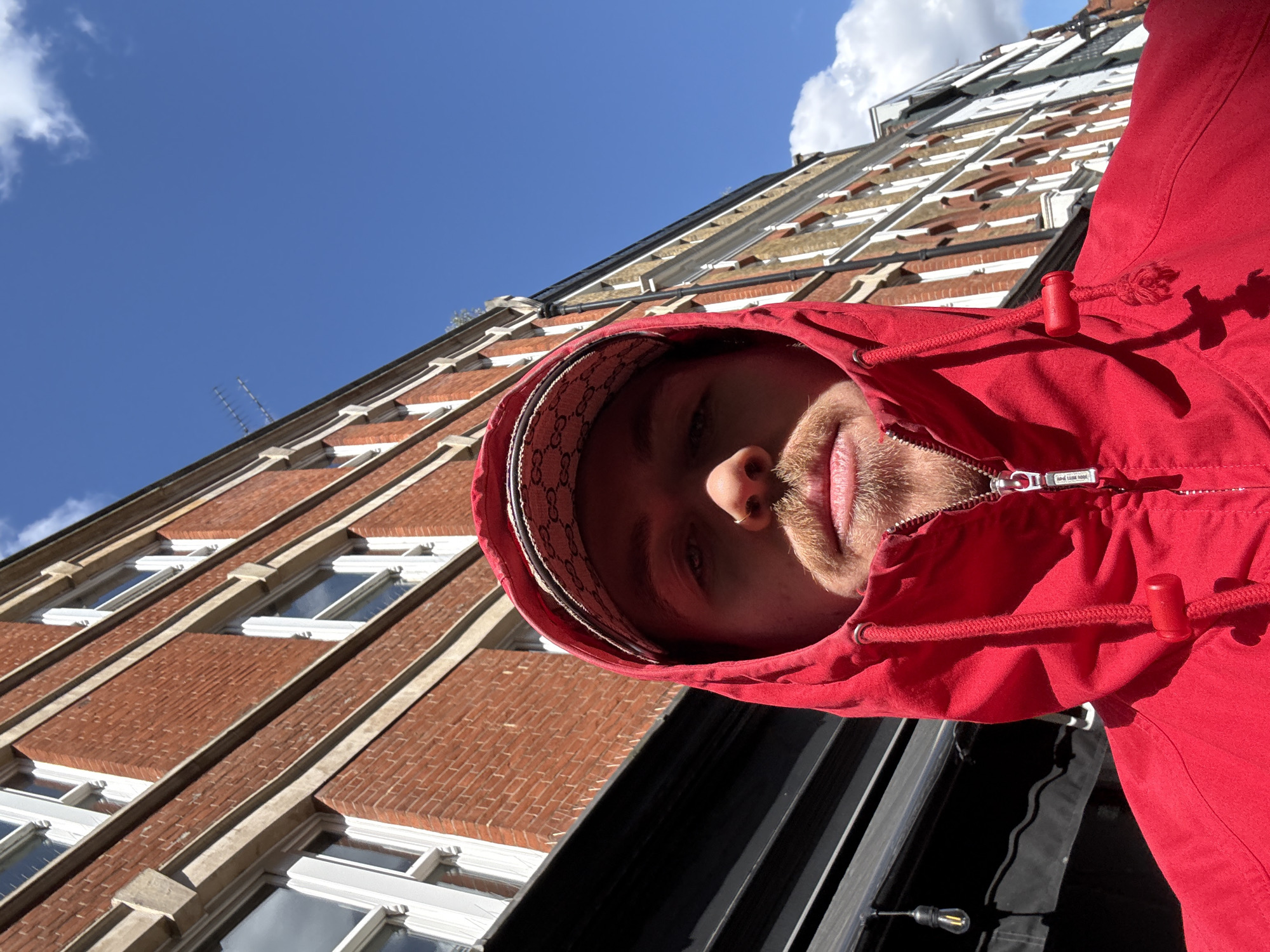 A low-angle selfie captures a person looking down towards the camera, wearing a bright red hooded jacket. Only the lower half of their face is visible within the hood, including their nose, mouth, and chin, framed by light-colored stubble or a short beard. Underneath the red hood, a cap with a repeating decorative pattern is worn, providing a distinctive trim. The jacket features a prominent silver zipper pull that reads "HUGO BOSS" and a red plastic toggle on a drawstring.
The background reveals a sunny urban setting, likely in London, United Kingdom, given the context. Above the person, a clear blue sky with a few fluffy white clouds is visible. To the left and behind them, a tall brick building with multiple floors stretches upwards, characterized by its reddish-brown bricks and rows of white-framed windows with stone lintels and sills. The perspective makes the buildings appear angled, adding to the dynamic composition. The scene suggests a bright, clear day.