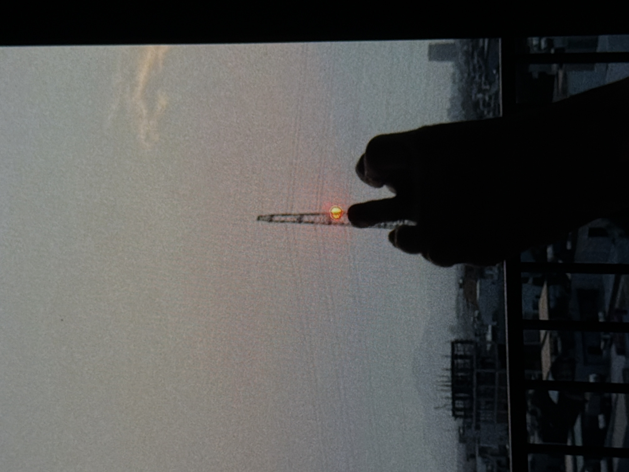 The image shows a cityscape with a muted, hazy sky, possibly taken in Đà Nẵng, Vietnam. A hand is reaching out, partially obscuring the view. In the distance, a construction crane is visible, appearing to be illuminated with a small yellow sun behind it. The cityscape includes buildings and structures, with a balcony railing visible in the foreground. The image appears to be taken through a screen or window.
