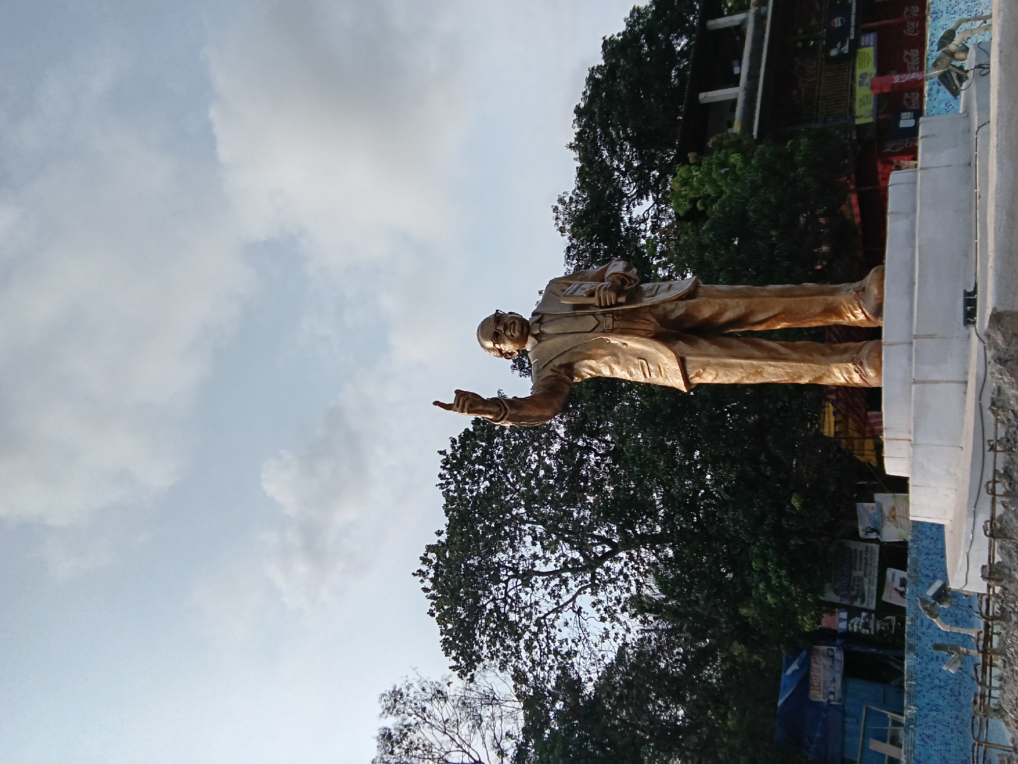 A low-angle shot captures a bronze statue of a man in a suit, likely Dr. B.R. Ambedkar, atop a white pedestal in Chopdem, India. The man wears glasses and holds a book in one hand, while pointing forward with the other. Behind the statue, dark green foliage fills the frame. The cloudy sky is visible in the upper portion of the shot. In the background, there are buildings with signage and advertisements. The base of the statue and the surrounding area appear to be under construction, as indicated by exposed rebar and unfinished mosaic work.
