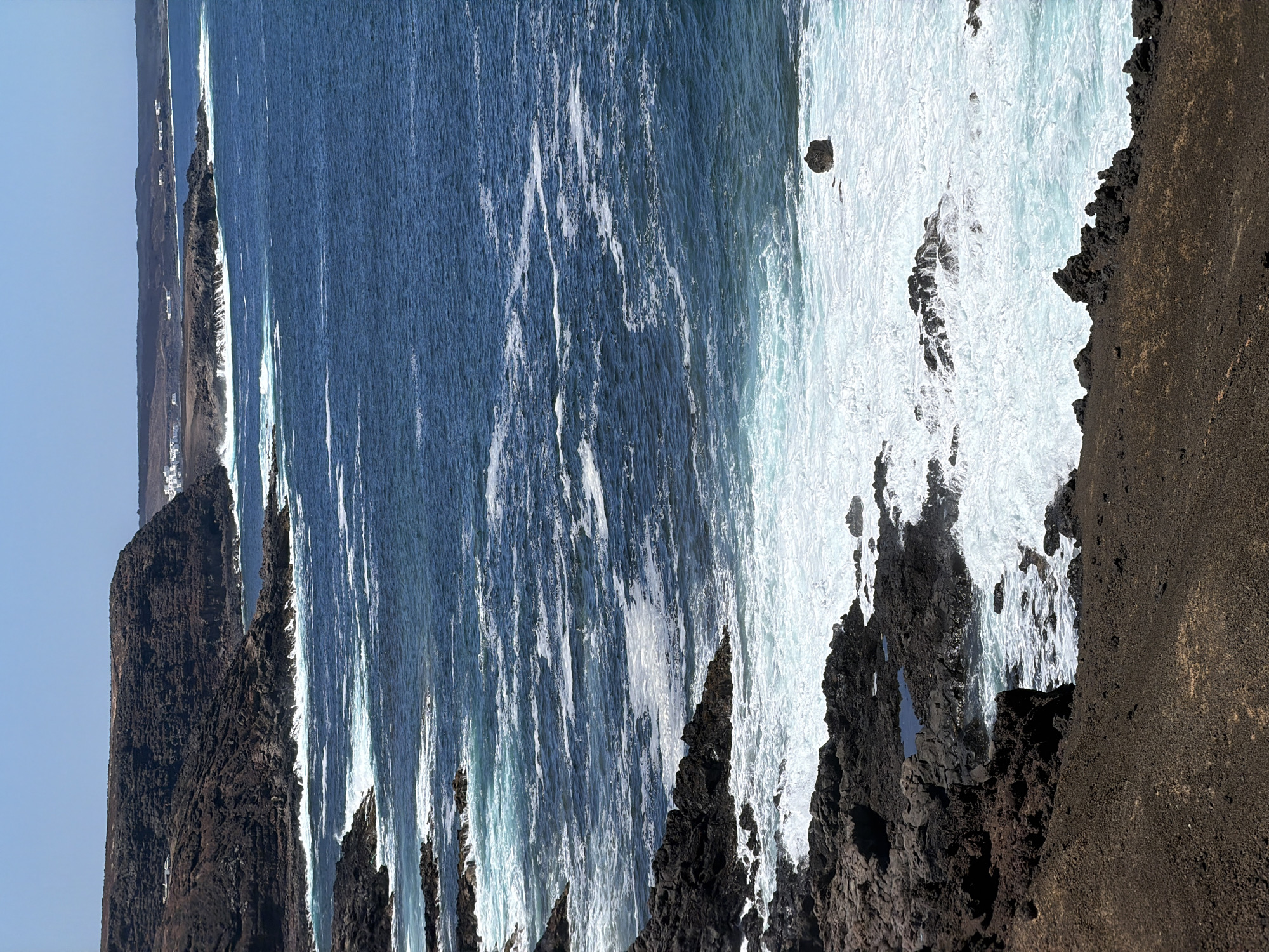 Waves crash against the jagged, dark volcanic coastline of Tinajo, Spain. The sky is a clear, pale blue. The water transitions from deep blue to frothy white where the waves meet the shore. The rocky terrain is rough and uneven.