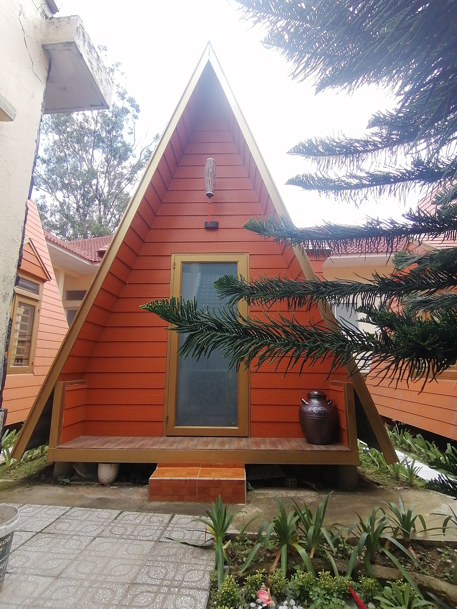 This image shows an A-frame cabin in Da Lat, Vietnam. The cabin is orange with a golden trim and has a dark brown door with a glass pane. A large, dark brown ceramic jar sits on the porch. The cabin is surrounded by lush greenery, including pine trees. In the foreground, there is a tiled pathway and some plants. The sky is overcast.